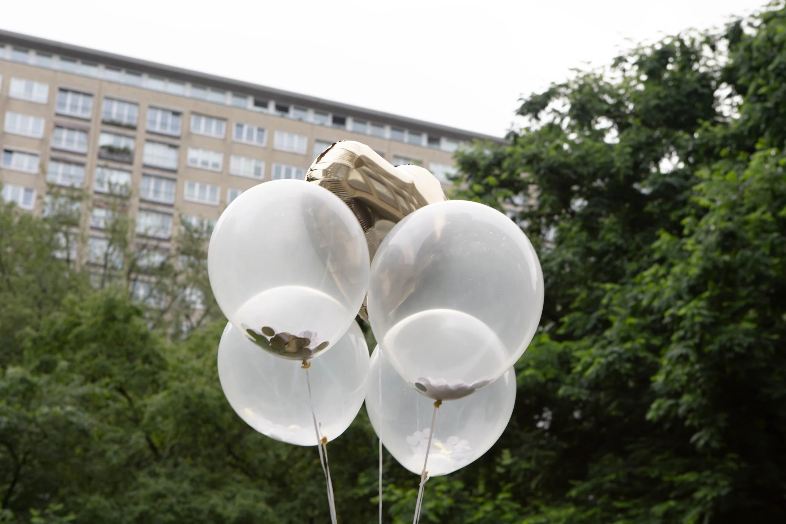 Mehrere durchsichtige Luftballons mit weißen Ballonpuffen im Freien, im Hintergrund ein Hochhaus und grüne Bäume.
