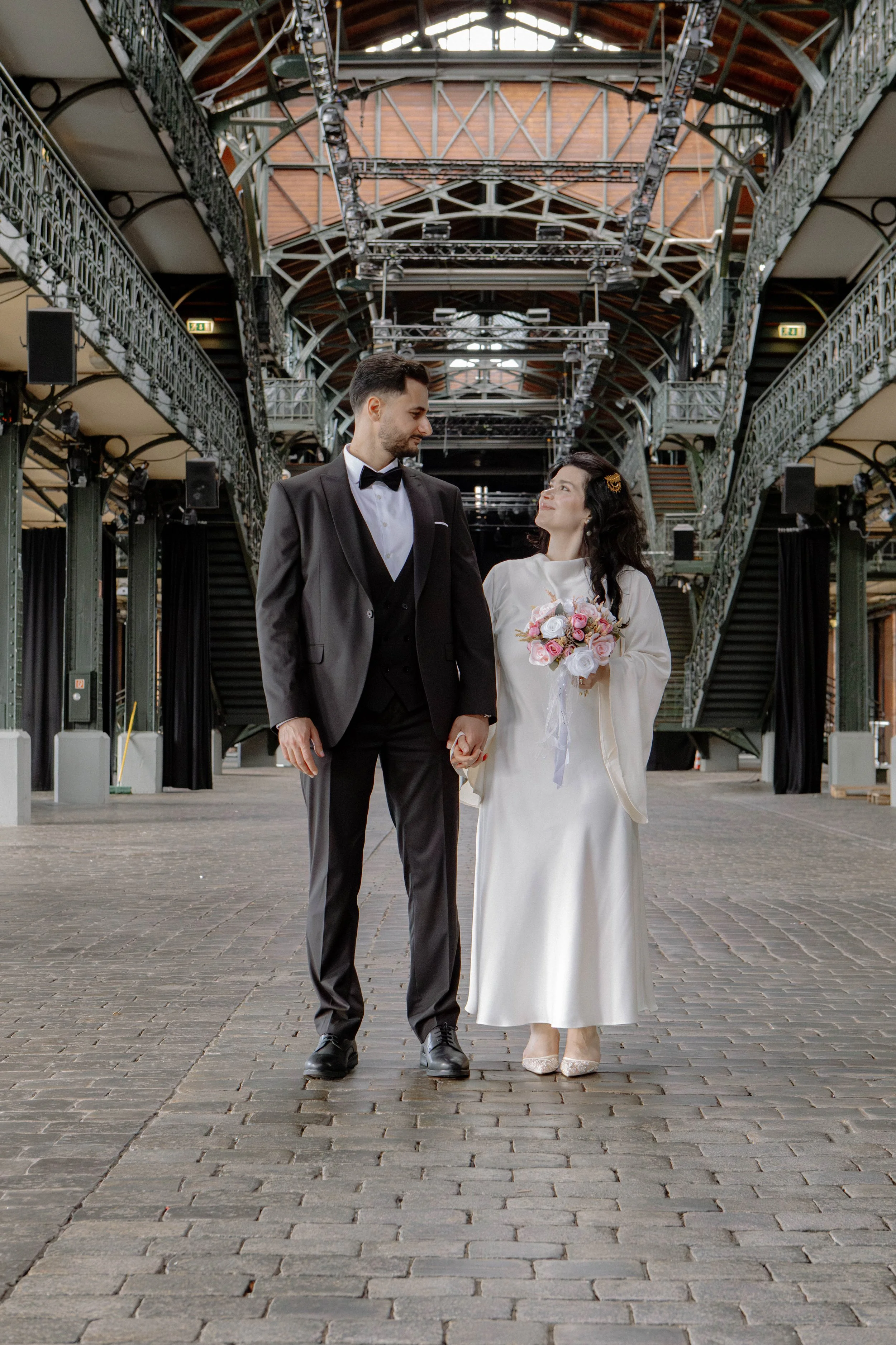 Ein Hochzeitsfoto mit einem verheirateten Paar, das miteinander spazieren geht. Sie sind auf einer gepflasterten Fläche in einem Gebäude mit einem hohen, bogenförmigen Dach. Der Mann trägt einen schwarzen Anzug und eine Fliege, die Frau trägt ein lan