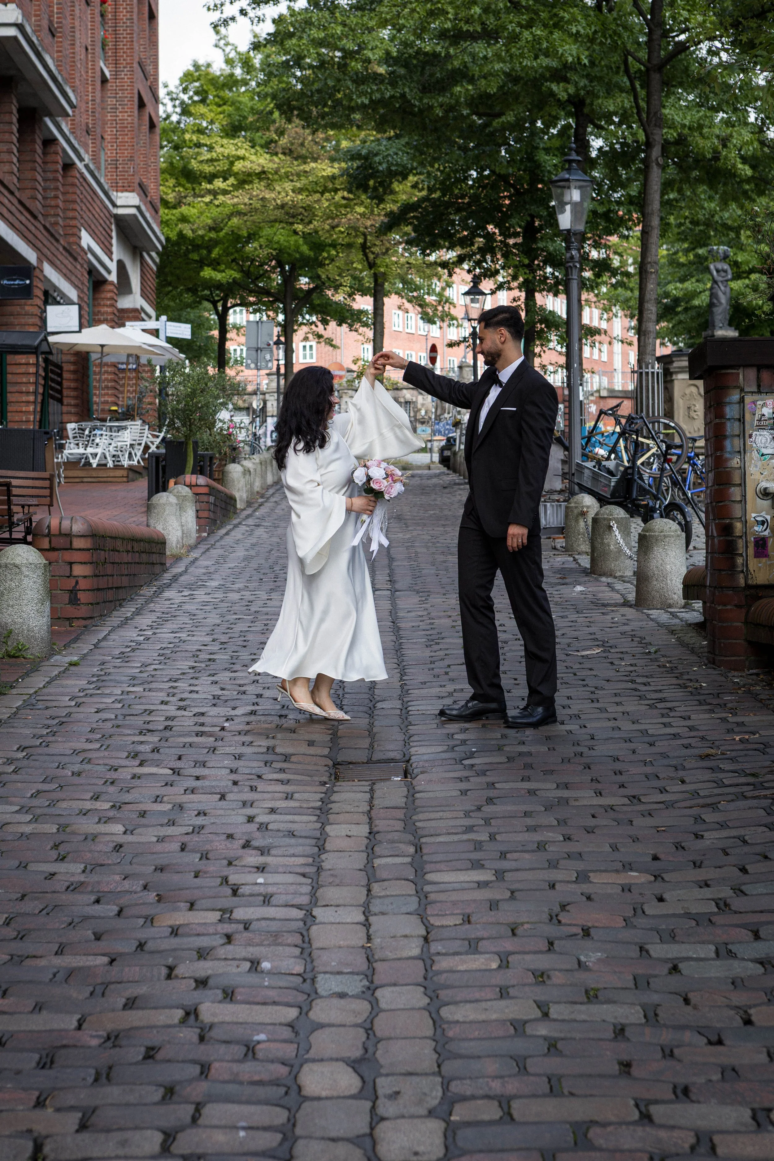 Ein Mann in einem schwarzen Anzug und eine Frau in einem weißen Kleid tanzen auf einer gepflasterten Straße. Die Frau hält einen Blumenstrauß, und der Mann überreicht ihr ein Geschenk, möglicherweise eine Hochzeitsfeier.