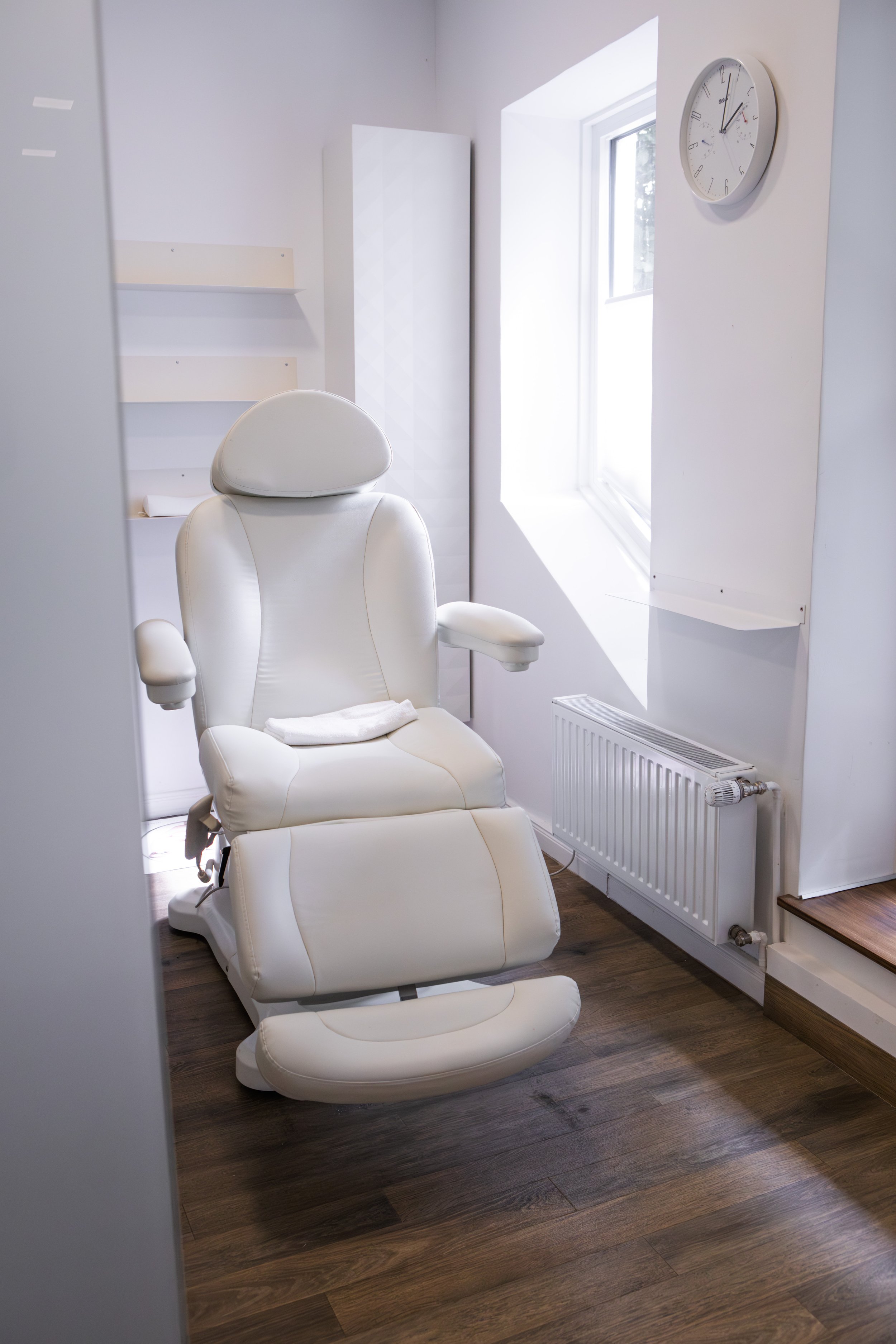 Ein weißer medizinischer Untersuchungstuhl in einem hellen, minimalistisch eingerichteten Raum, mit Fenster und Uhr an der Wand.