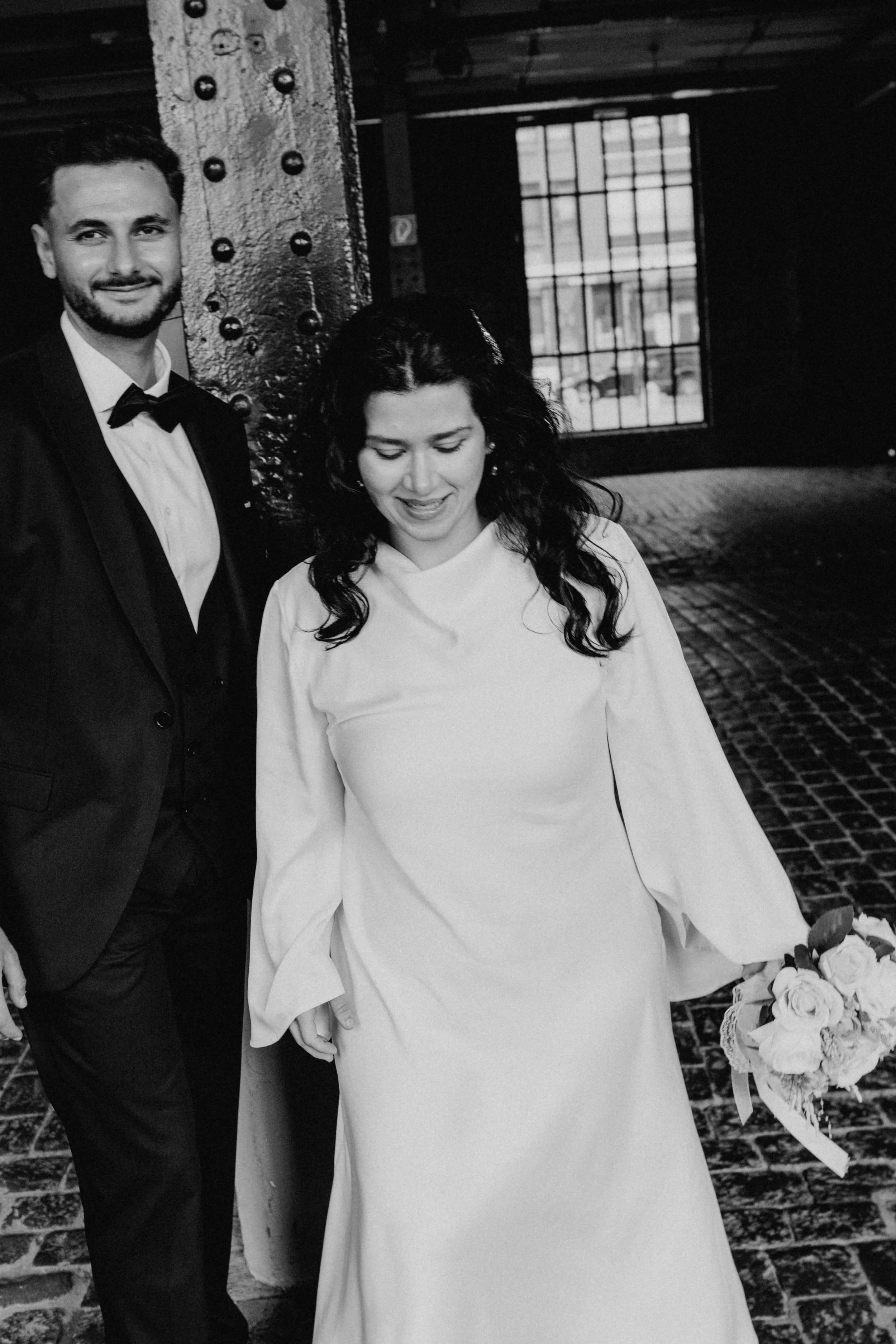 Ein Mann im schwarzen Anzug mit Fliege und eine Frau im weißen Brautkleid, die einen Blumenstrauß hält, in einer dunklen, industrialisierten Umgebung mit großen Fenstern.