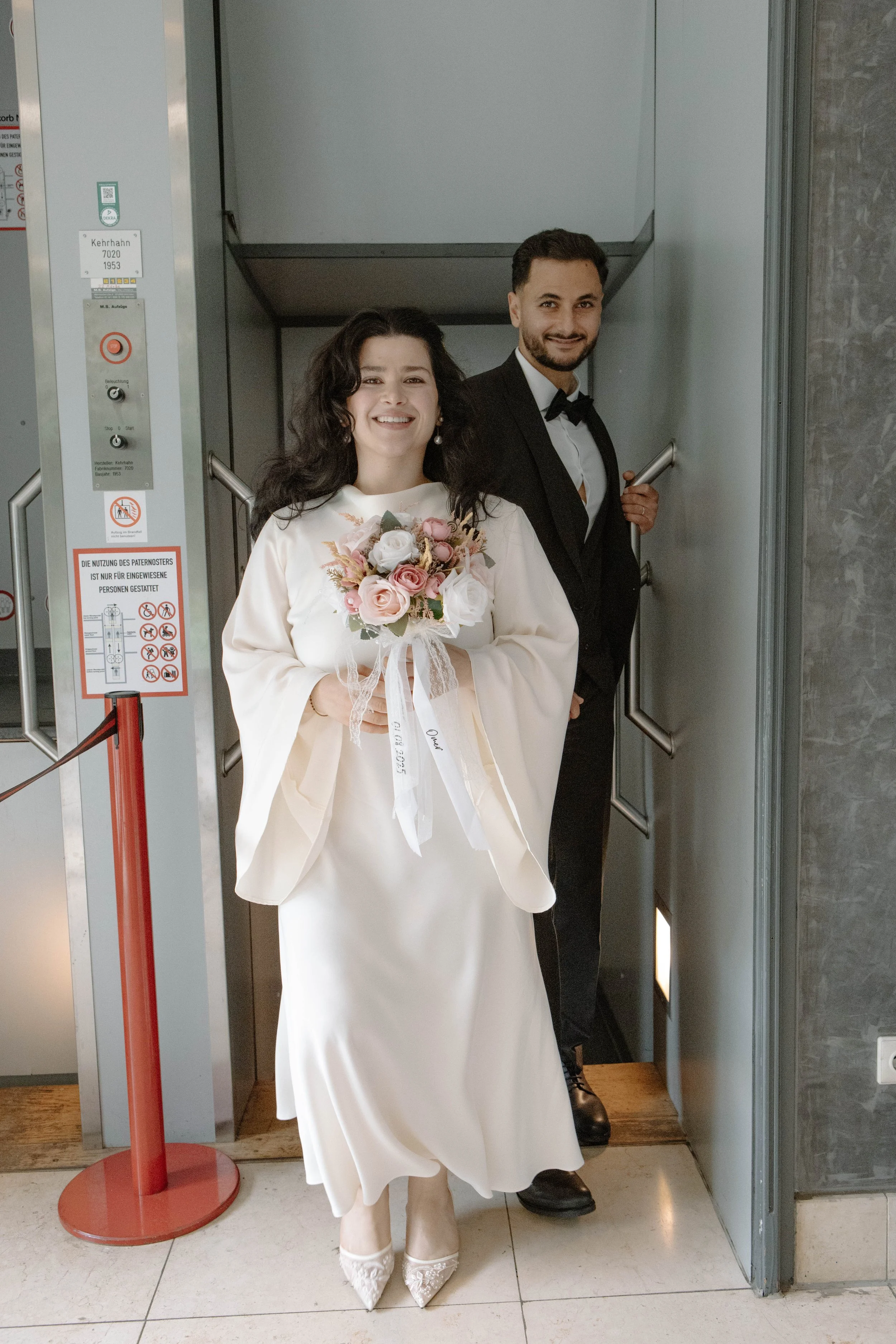 Ein Hochzeiterpaar steht in einem Aufzug, der Bräutigam in einem Anzug und die Braut in einem weißen Kleid mit einem Blumenstrauß.