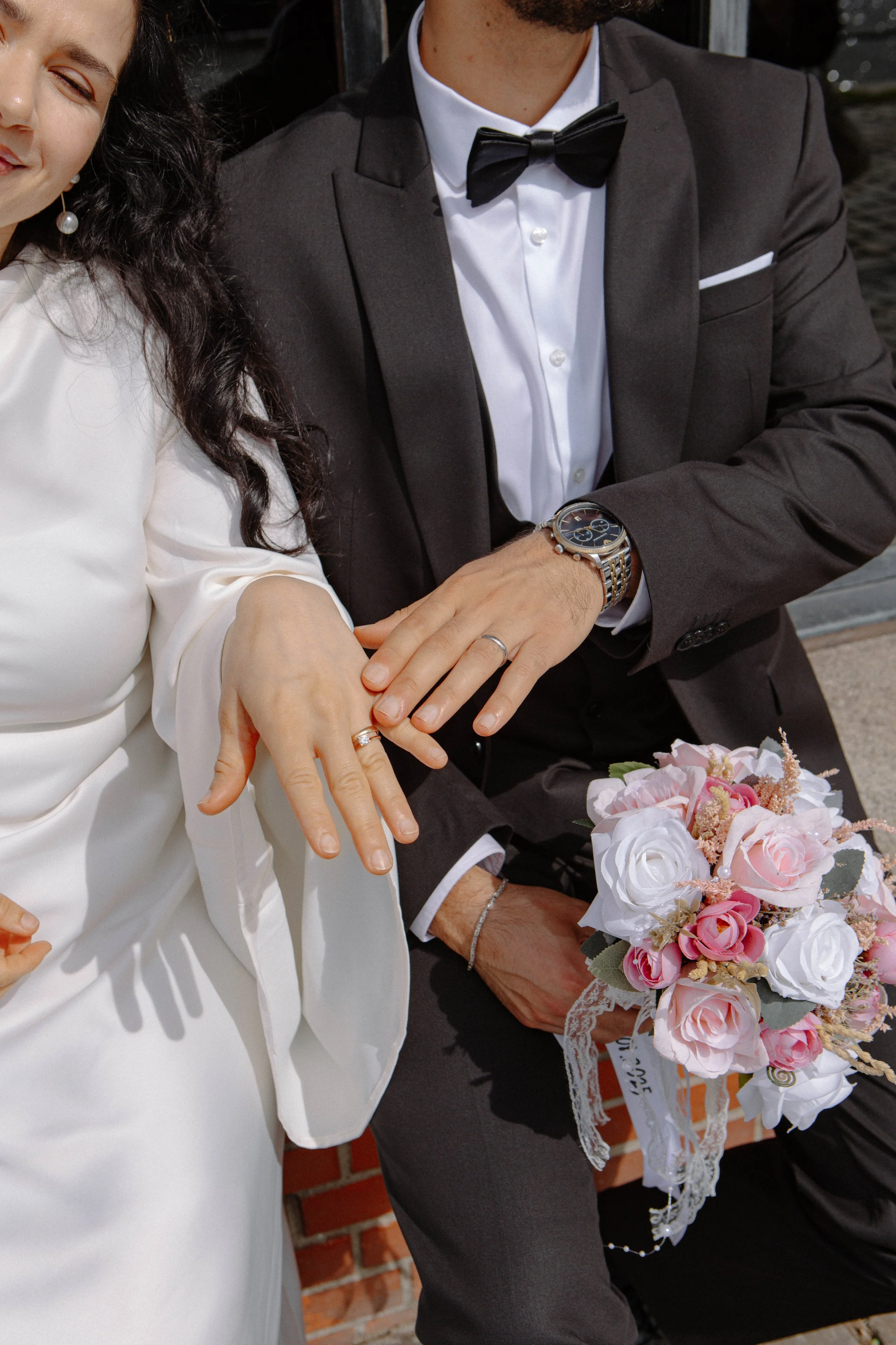Ein Brautpaar zeigt ihre Eheringe, die Braut trägt ein weißes Kleid und der Bräutigam einen schwarzen Anzug mit Fliege, sie halten ihre Hände über einem Blumenstrauß mit rosa und weißen Rosen.