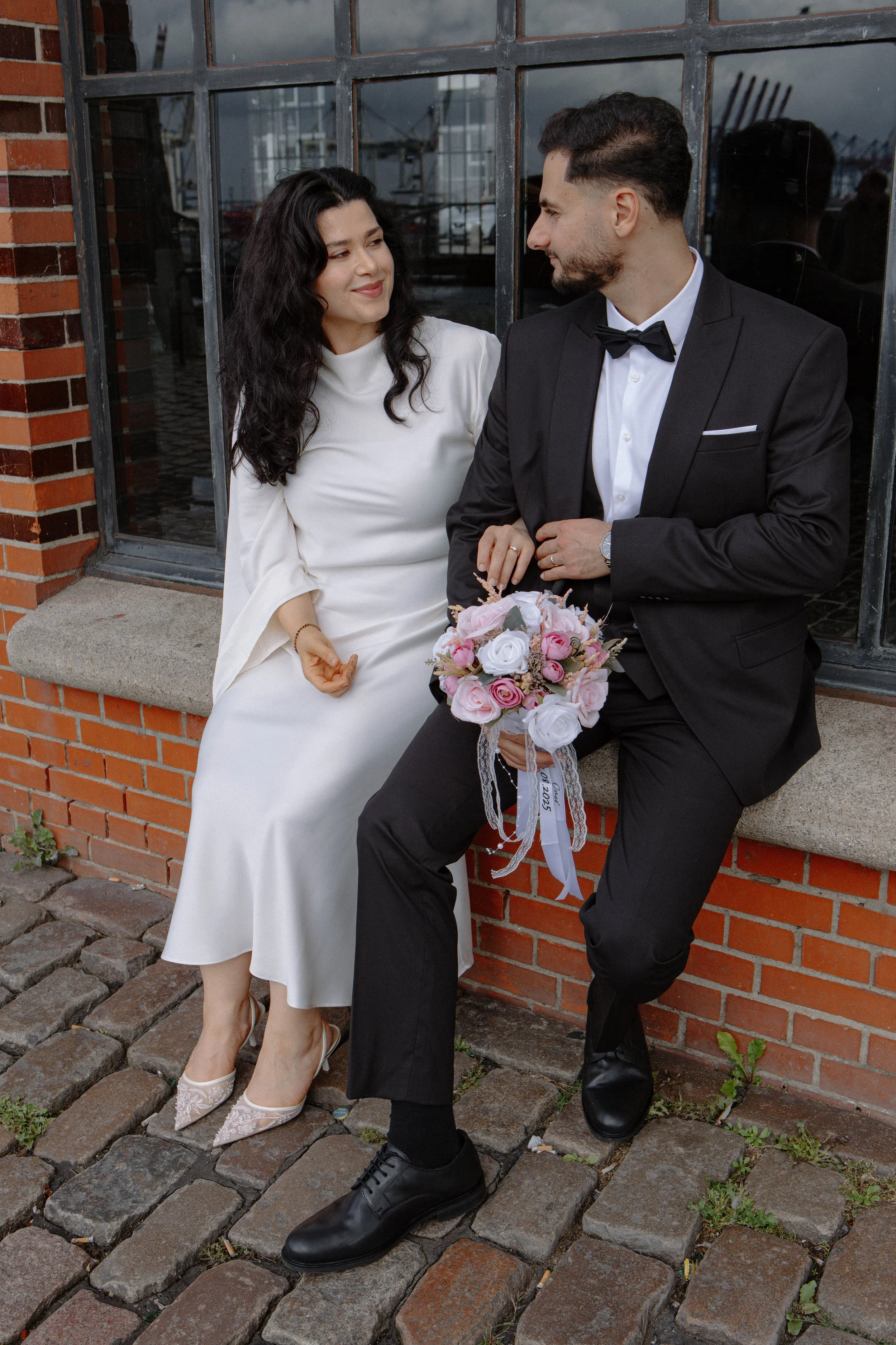 Ein Brautpaar sitzt vor einem Backsteinmauer und blickt sich an, die Frau trägt ein weißes Hochzeitskleid und high heels, der Mann trägt einen schwarzen Anzug mit Fliege und hält einen Brautstrauß aus rosa und weißen Blumen.