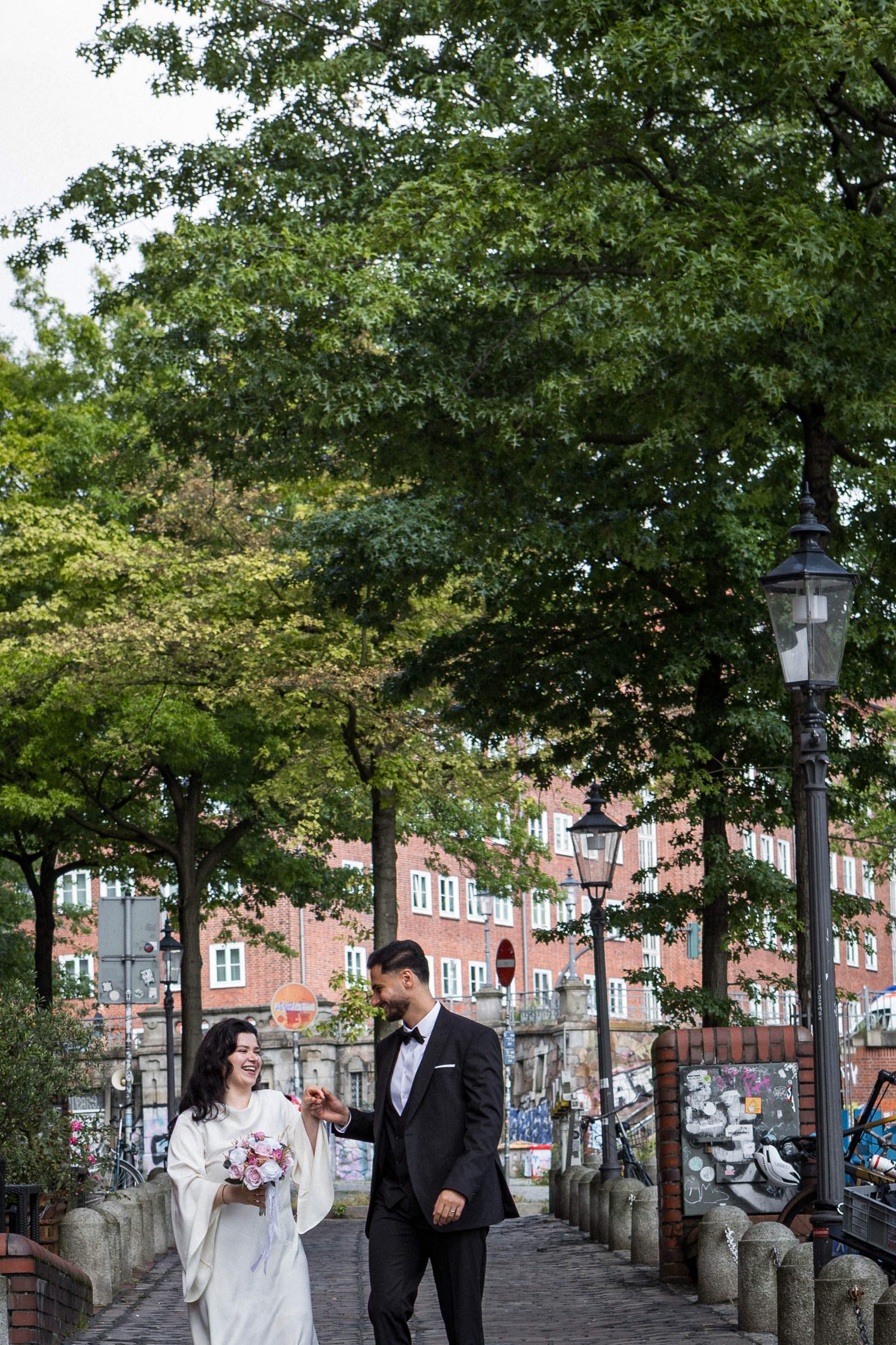 Ein glückliches Paar, die Braut in einem weißen Kleid und der Bräutigam in einem schwarzen Anzug mit Fliege, gehen auf einem Spazierweg unter Bäumen in einer Stadt, lachen und halten Händchen, die Braut trägt einen Blumenstrauß.