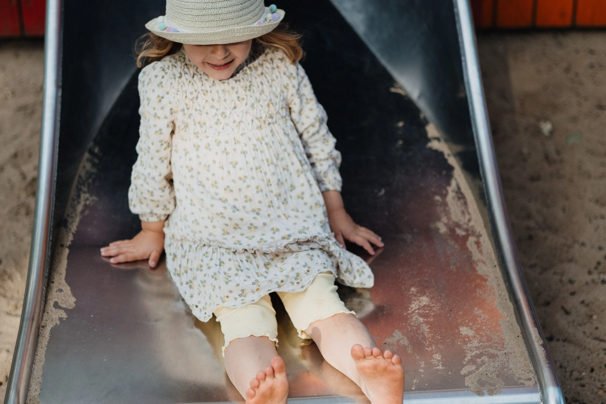 Ein lachendes Mädchen in einem weißen Kleid mit kleinen Blumen, das auf einer Rutschbahn im Sand spielt.
