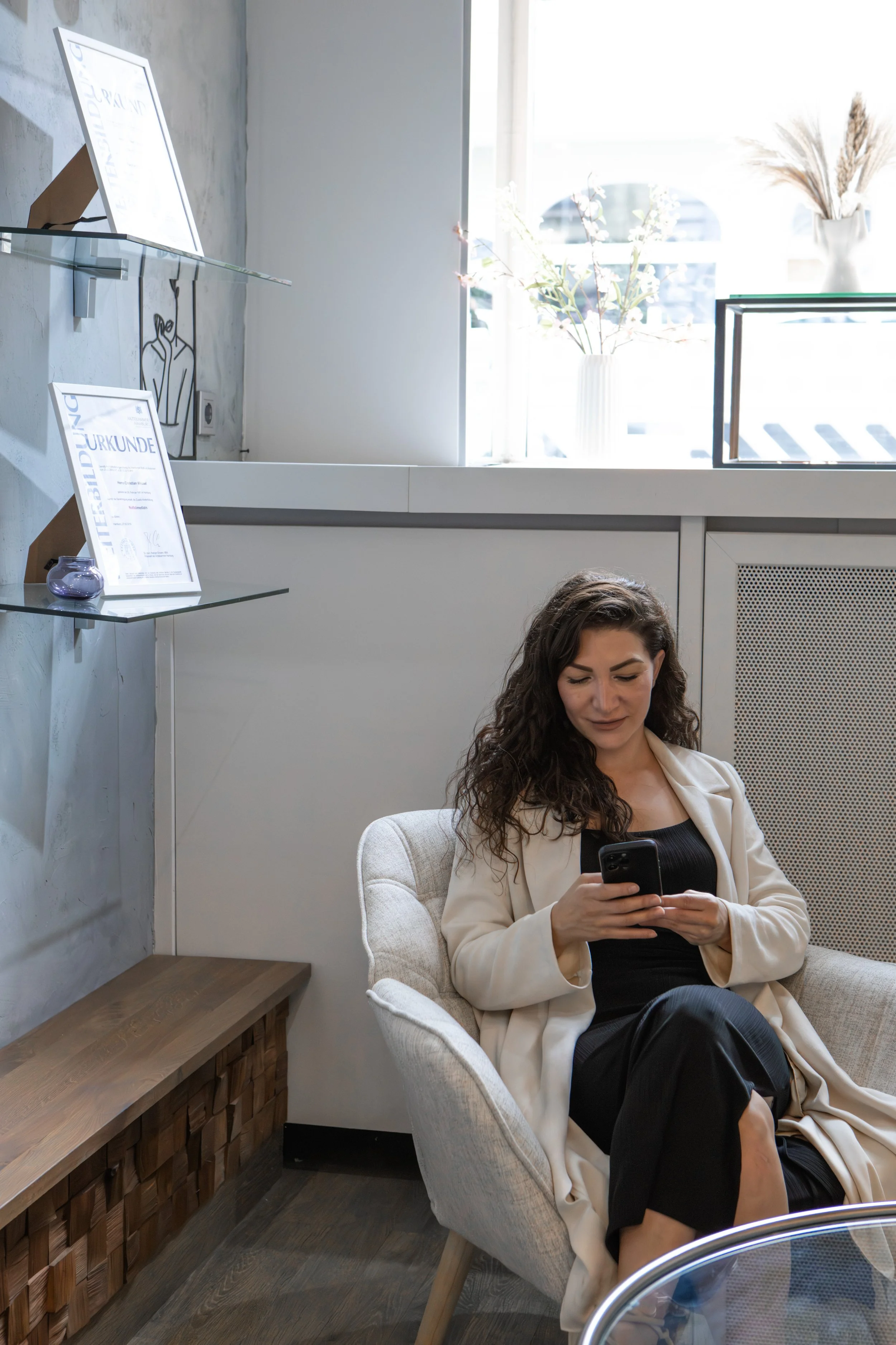 Eine Frau mit dunklen, lockigen Haaren sitzt in einem hellen Raum auf einem beige Couch und schaut auf ihr Smartphone.