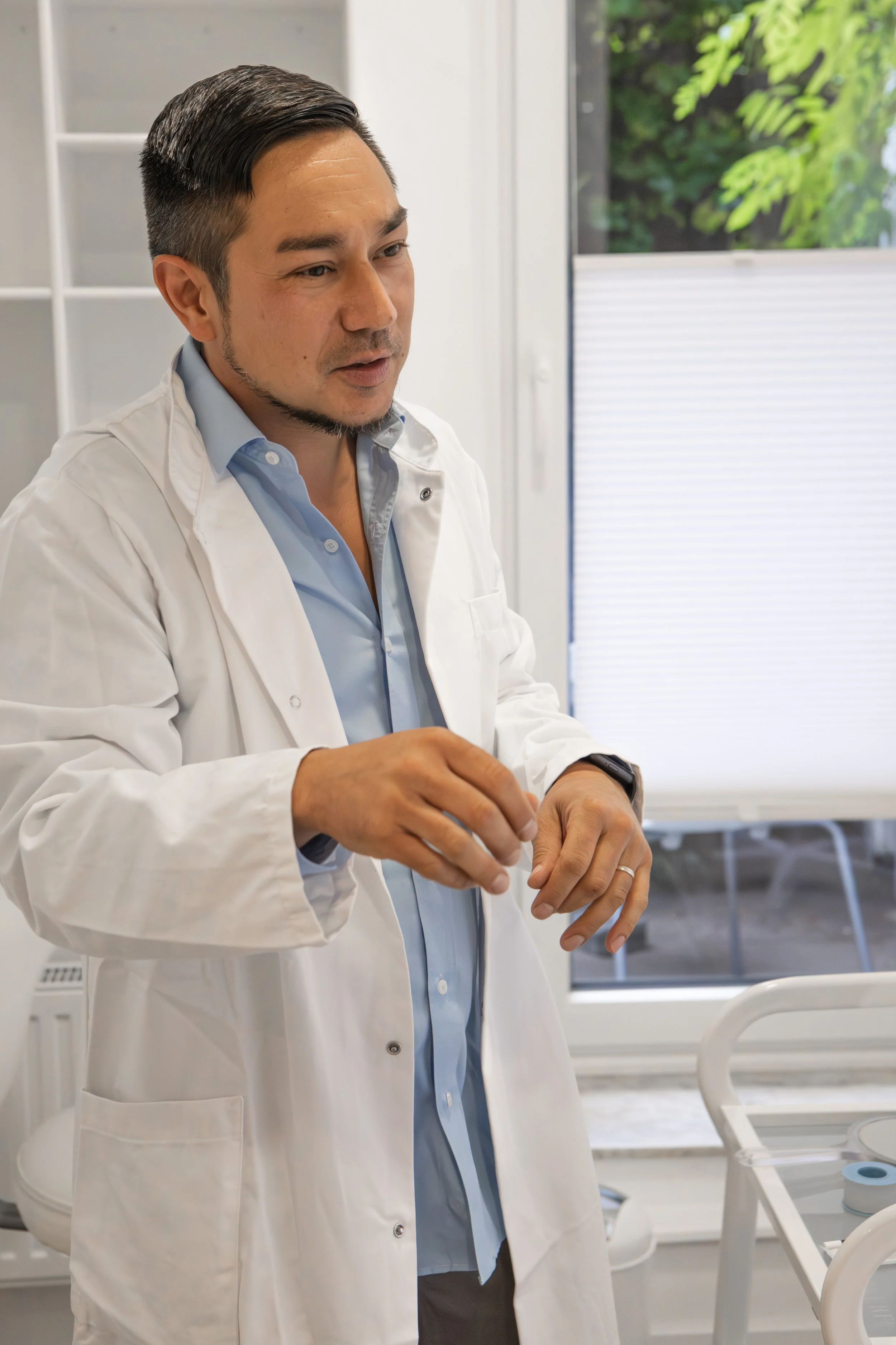 Ein Mann in weißem Arztkittel erklärt etwas in einem Krankenhauszimmer mit Betten und medizinischer Ausrüstung.