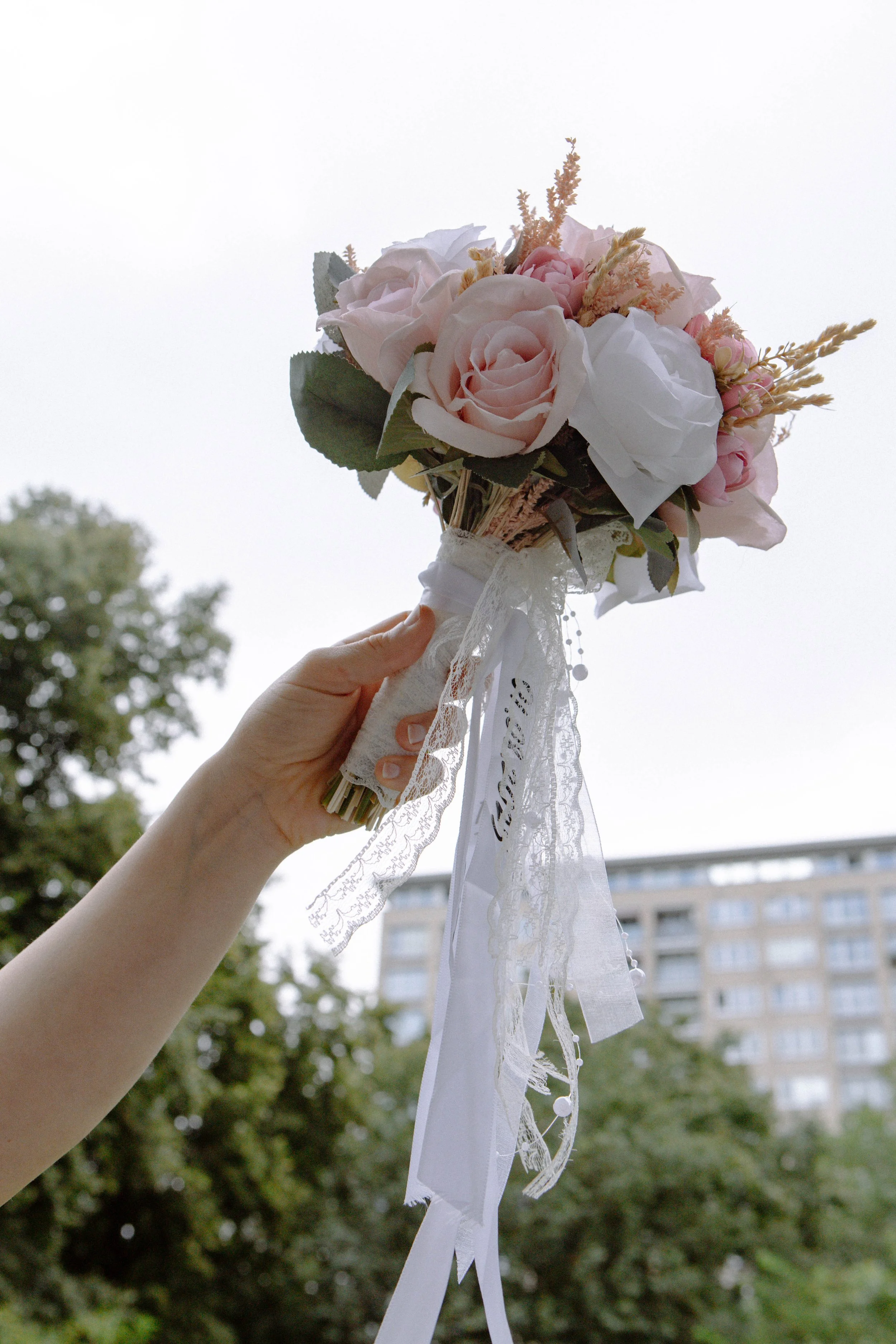 Eine Hand hält einen Brautstrauß aus rosa und weißen Rosen mit Lianen und Spitzenbändern, vor dem Hintergrund eines Stadtgebäudes und Bäumen.