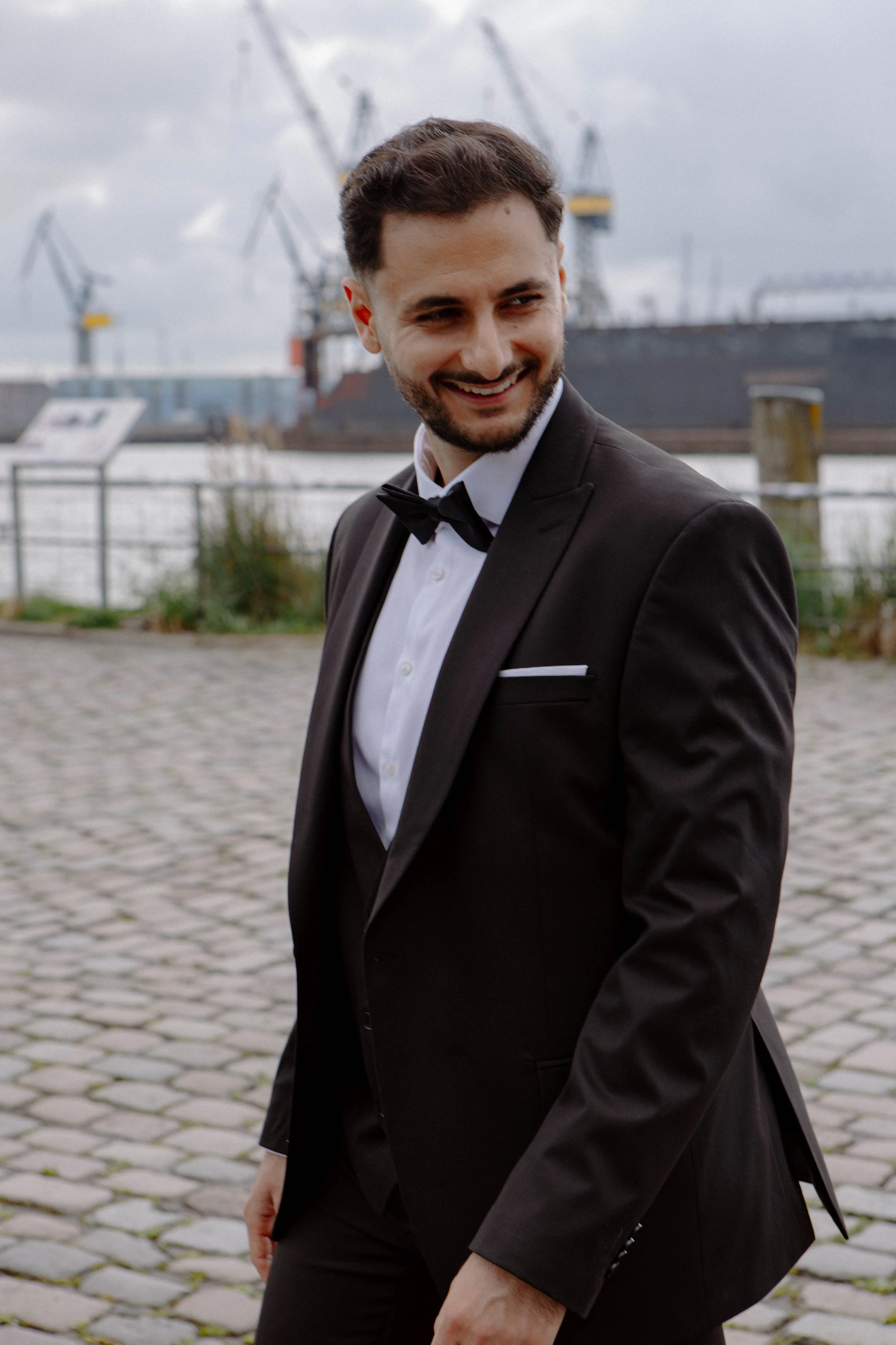 Junger Mann in formeller schwarzer Anzug mit Fliege, lächelnd an einer Hafengegend mit Krantoren im Hintergrund.