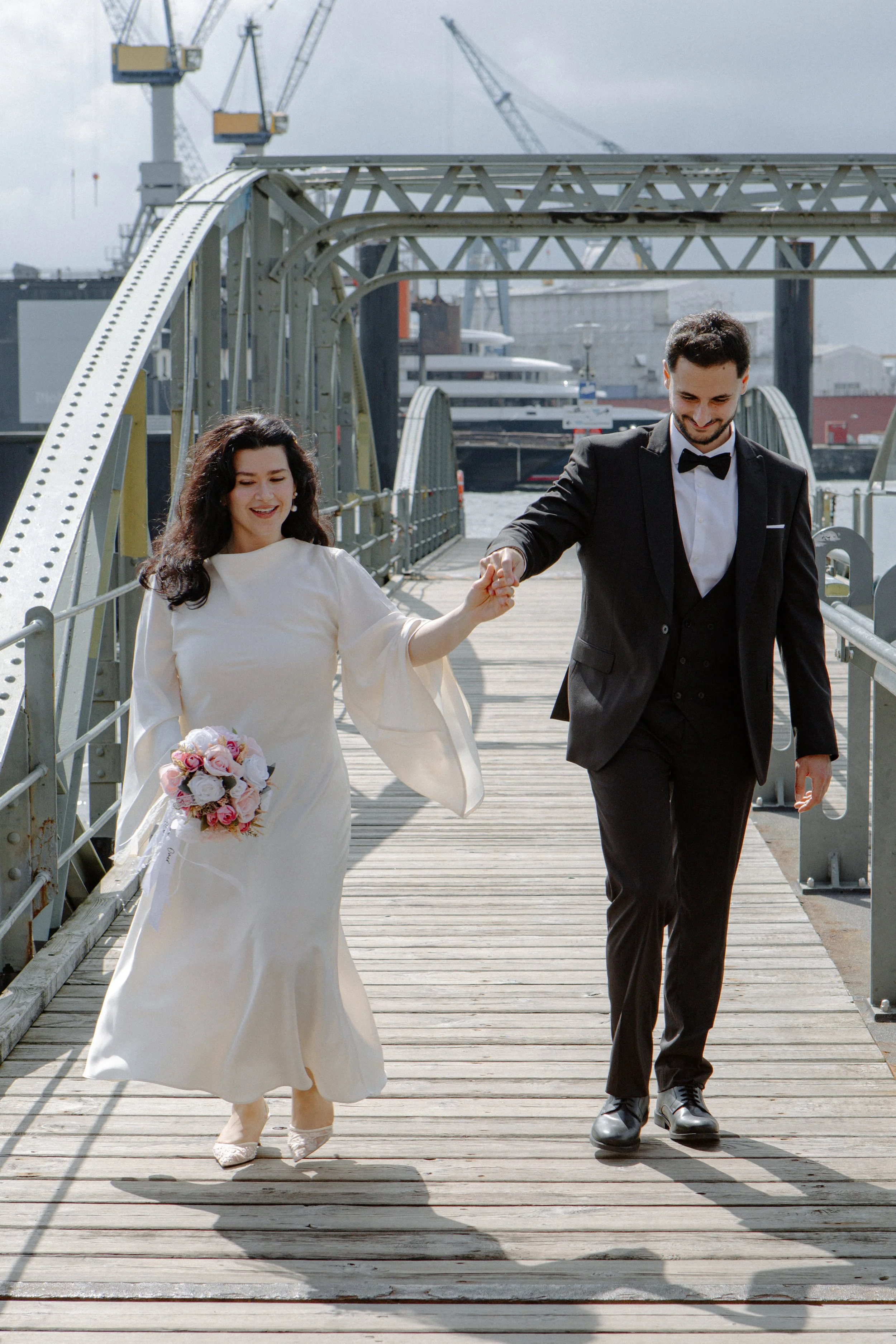 Ein frisch verheiratetes Paar, Frau in einem weißen Brautkleid mit Blumenstrauß, Mann in einem schwarzen Anzug mit Fliege, hält sich an der Hand und geht auf einer Holzbrücke am Wasser, im Hintergrund sind Kräne und Schiffe sichtbar.