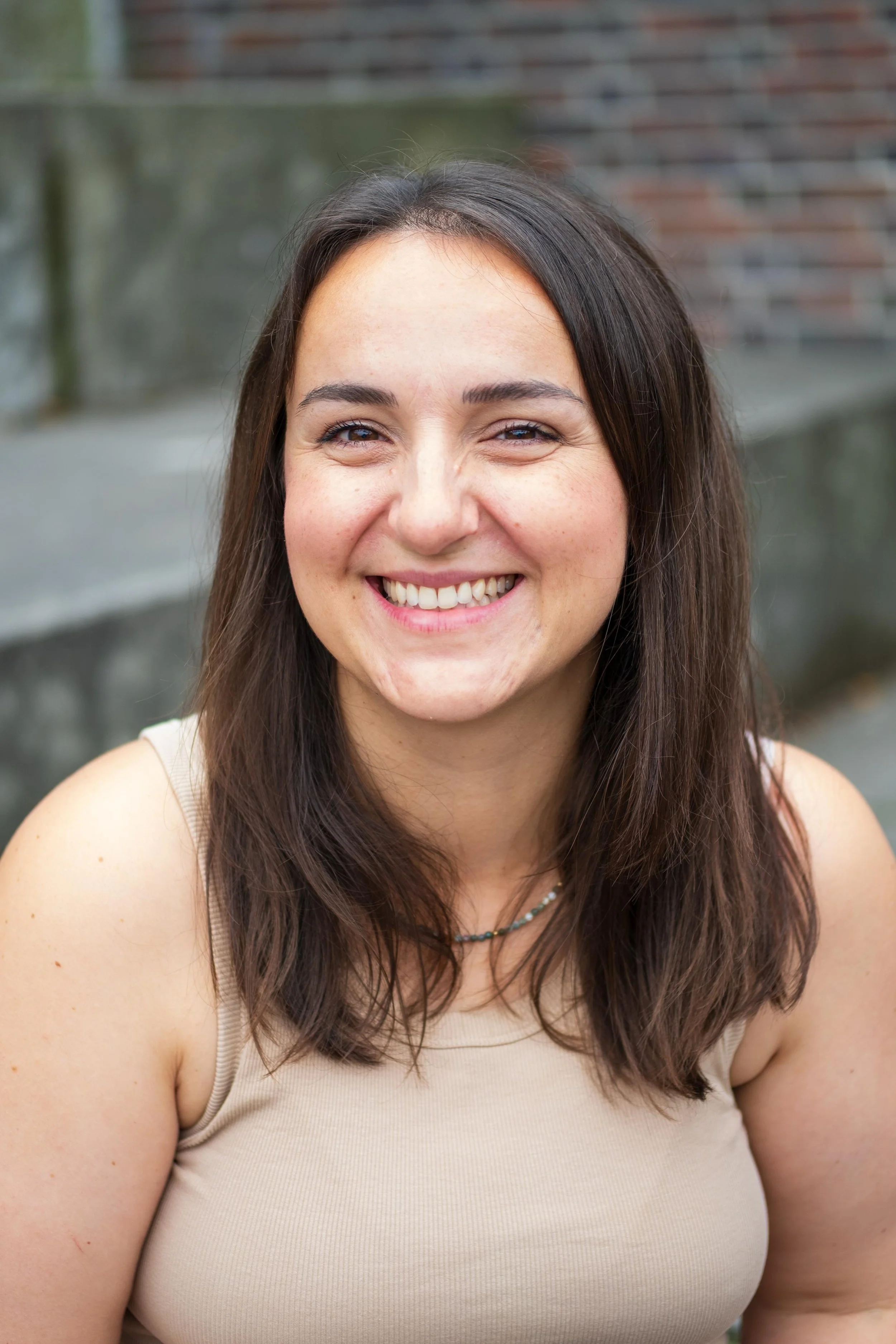 Junge Frau mit dunklen, schulterlangen Haaren, lächelnd, draußen vor einer Steinmauer, Träger eines beigen Tops und einer Halskette.