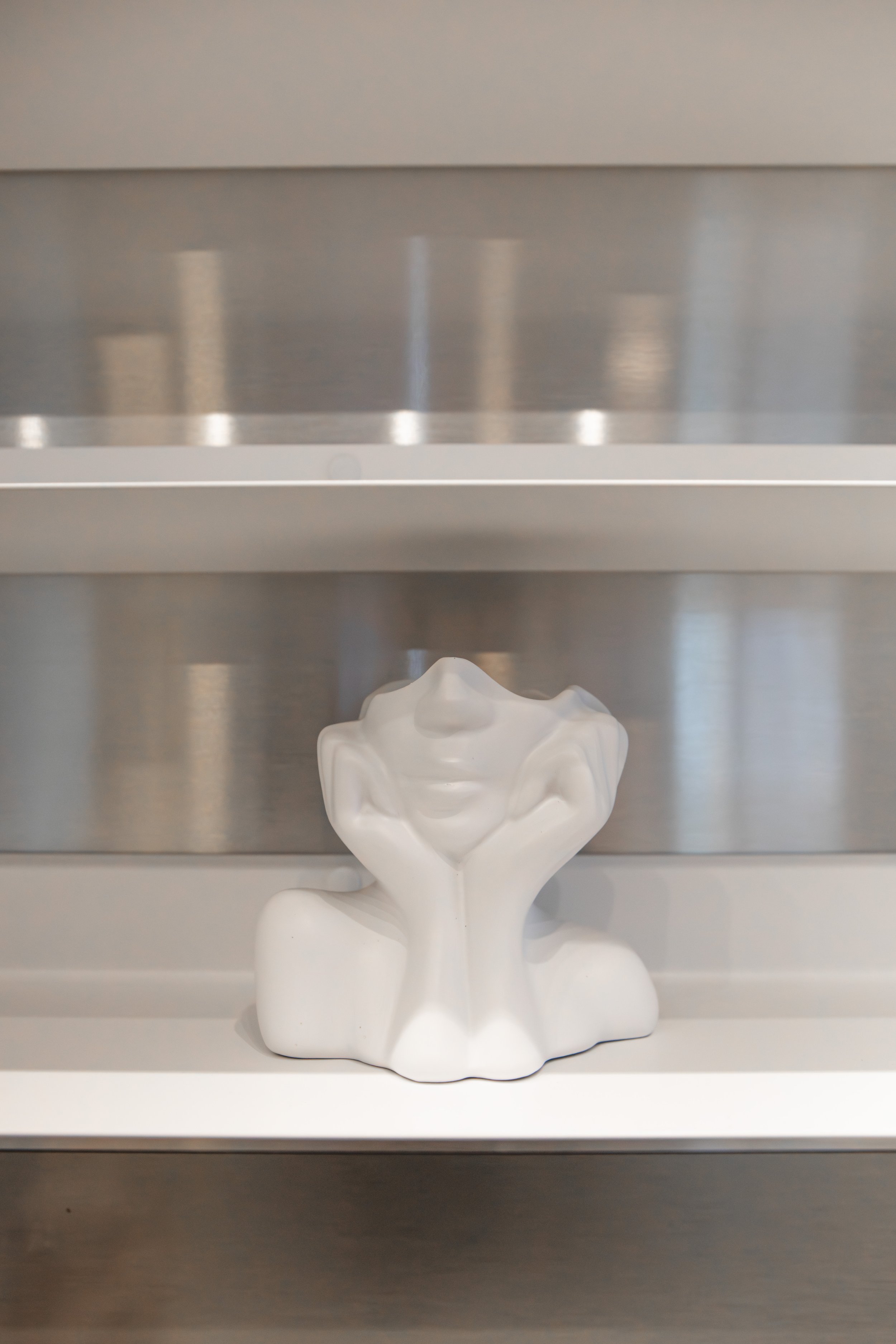 Weiße Skulptur eines Menschen, der seinen Kopf in die Hände stützt, auf einer weißen Oberfläche, im Hintergrund eine metallene Wand.