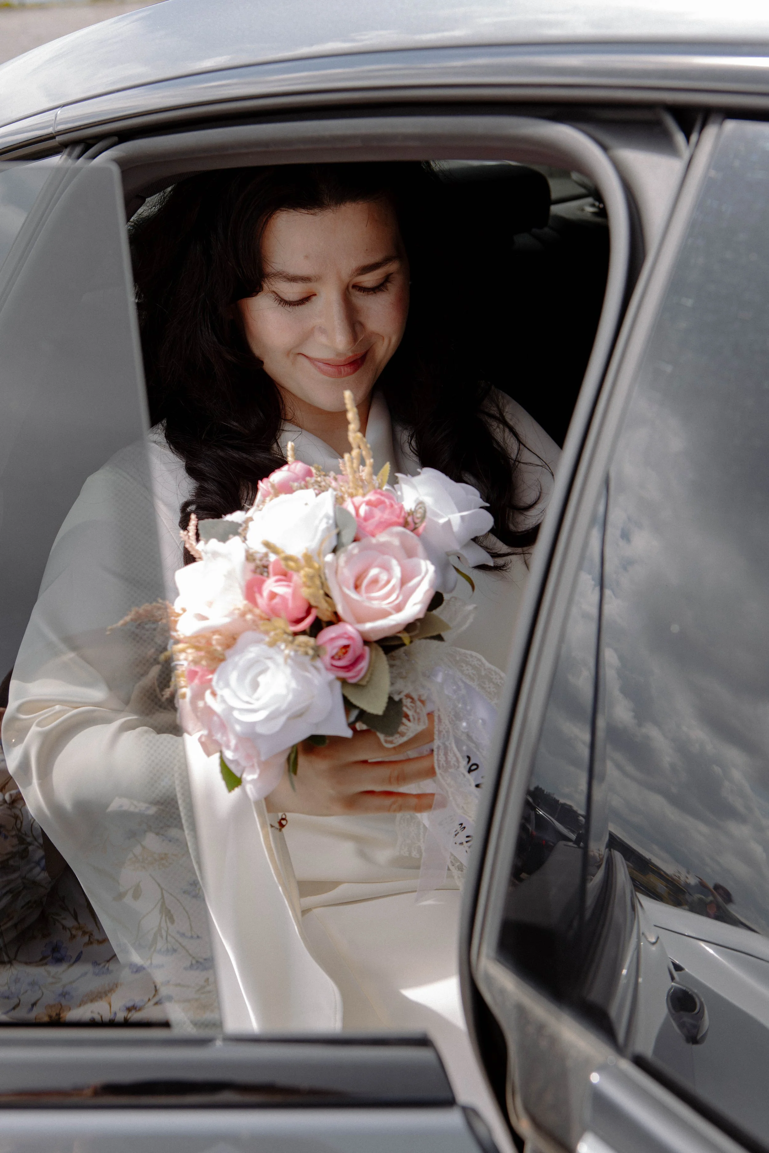 Eine Frau in einem weißen Brautkleid sitzt in einem Auto und hält einen Blumenstrauß aus rosa und weißen Blumen. Sie lächelt und schaut nach unten.