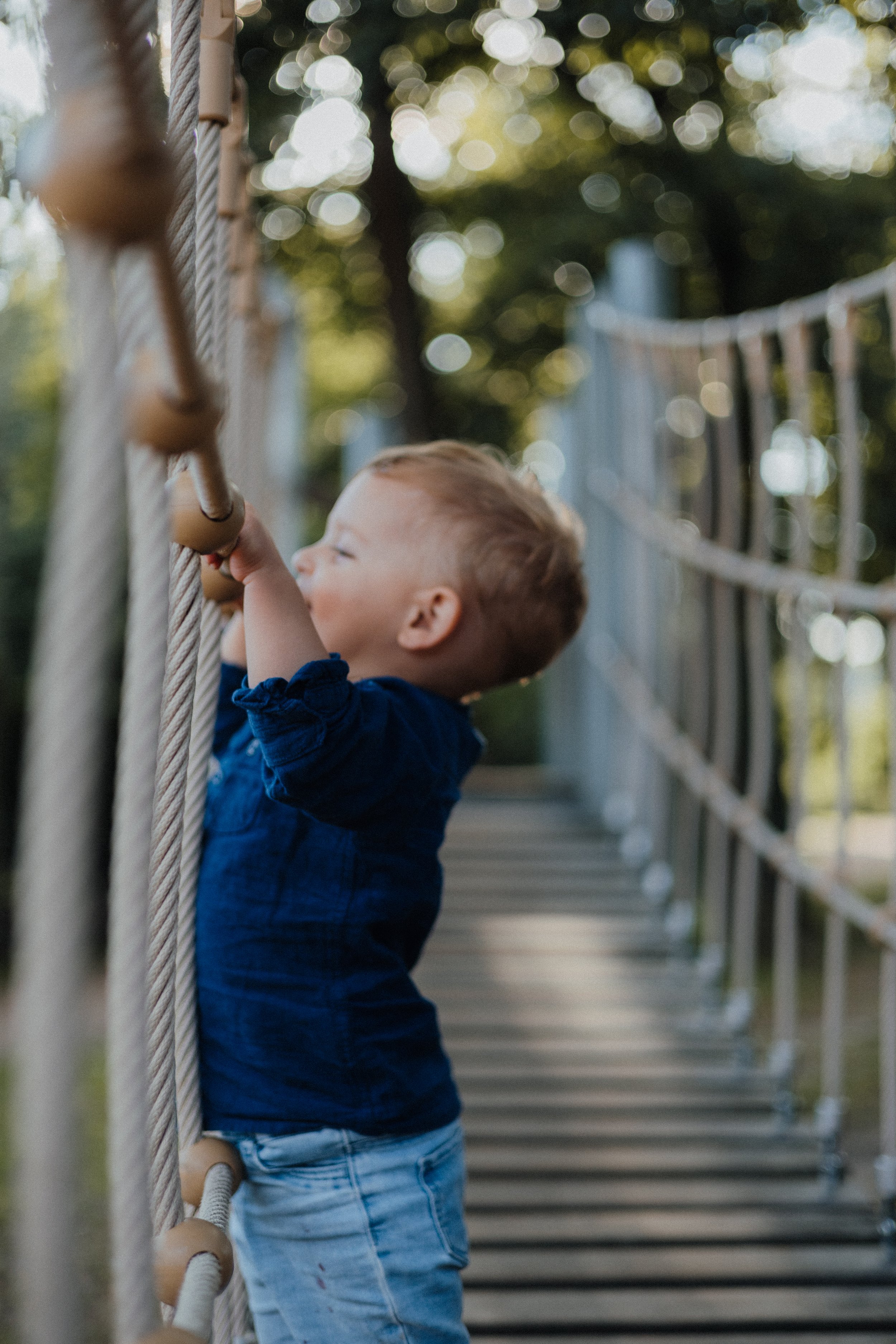 Junge spielt auf einer Hängebrücke im Freien, umgeben von Bäumen und Sonnenlicht.