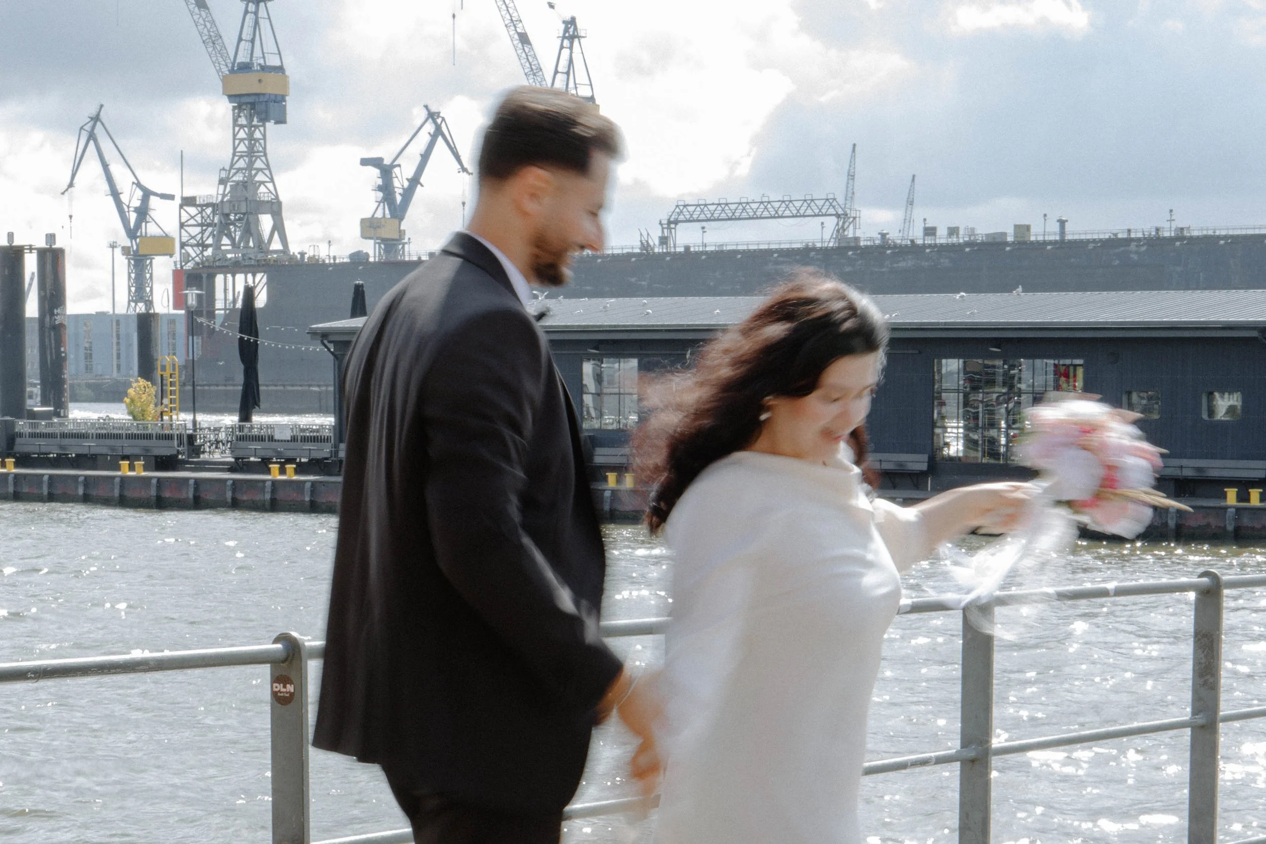 Ein Mann und eine Frau am Wasser, die Frau freut sich und hält einen Blumenstrauß, im Hintergrund sind Kräne und ein Industriehafen sichtbar.