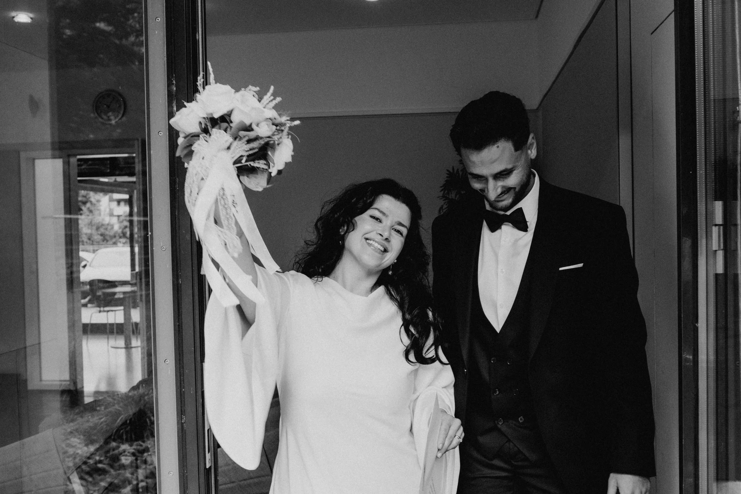 Ein glückliches Brautpaar freut sich, im Hochzeitskleid und im Anzug, beim Betreten eines Gebäudes, mit der Braut, die ein Blumenstrauß hochhält.