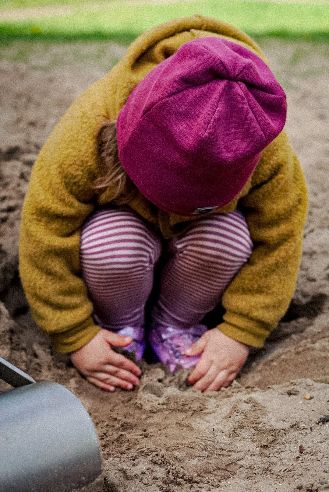 Ein Kind im grünlichen Sand spielt, trägt eine rosa Mütze und gestreifte Leggings, es kriecht im Sand.