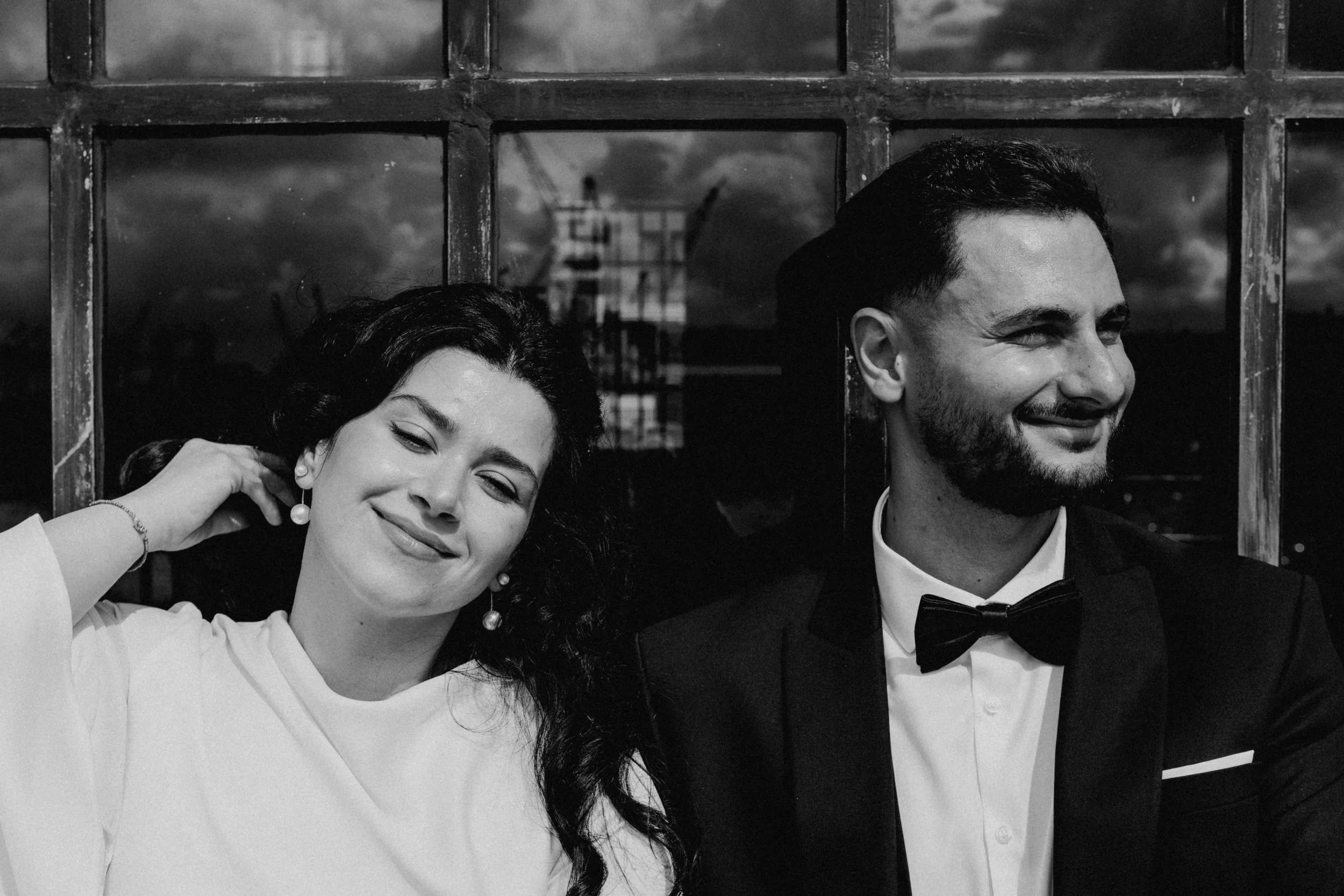 Ein Mann und eine Frau in formeller Kleidung, lächelnd, schwarz-weiß Foto, im Hintergrund eine Fensterfront mit Sicht auf einen Himmel mit Wolken.