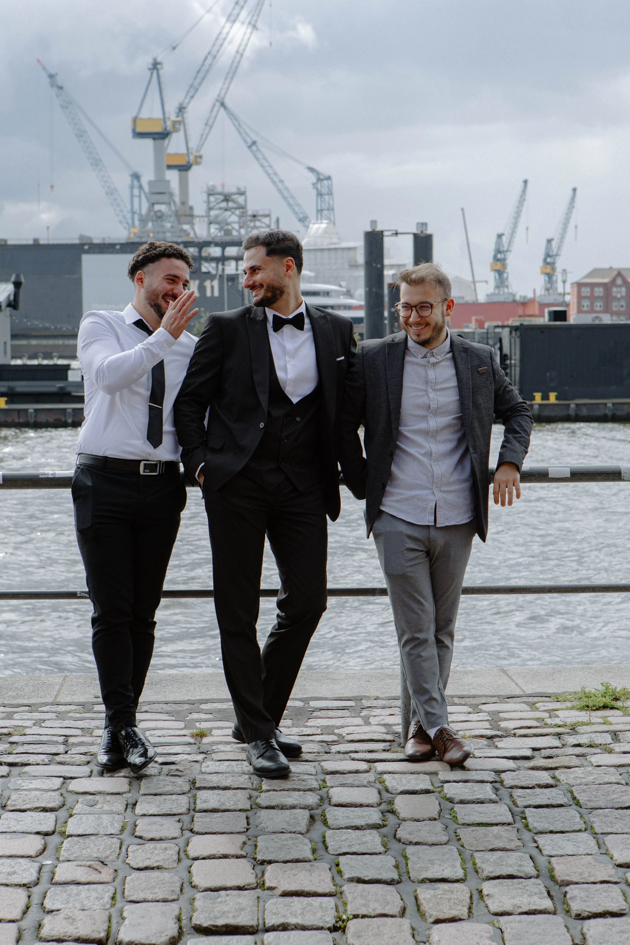 Drei Männer in Anzügen gehen lachend am Wasser entlang, im Hintergrund sind Kräne und ein Schiff in einem Hafen sichtbar.