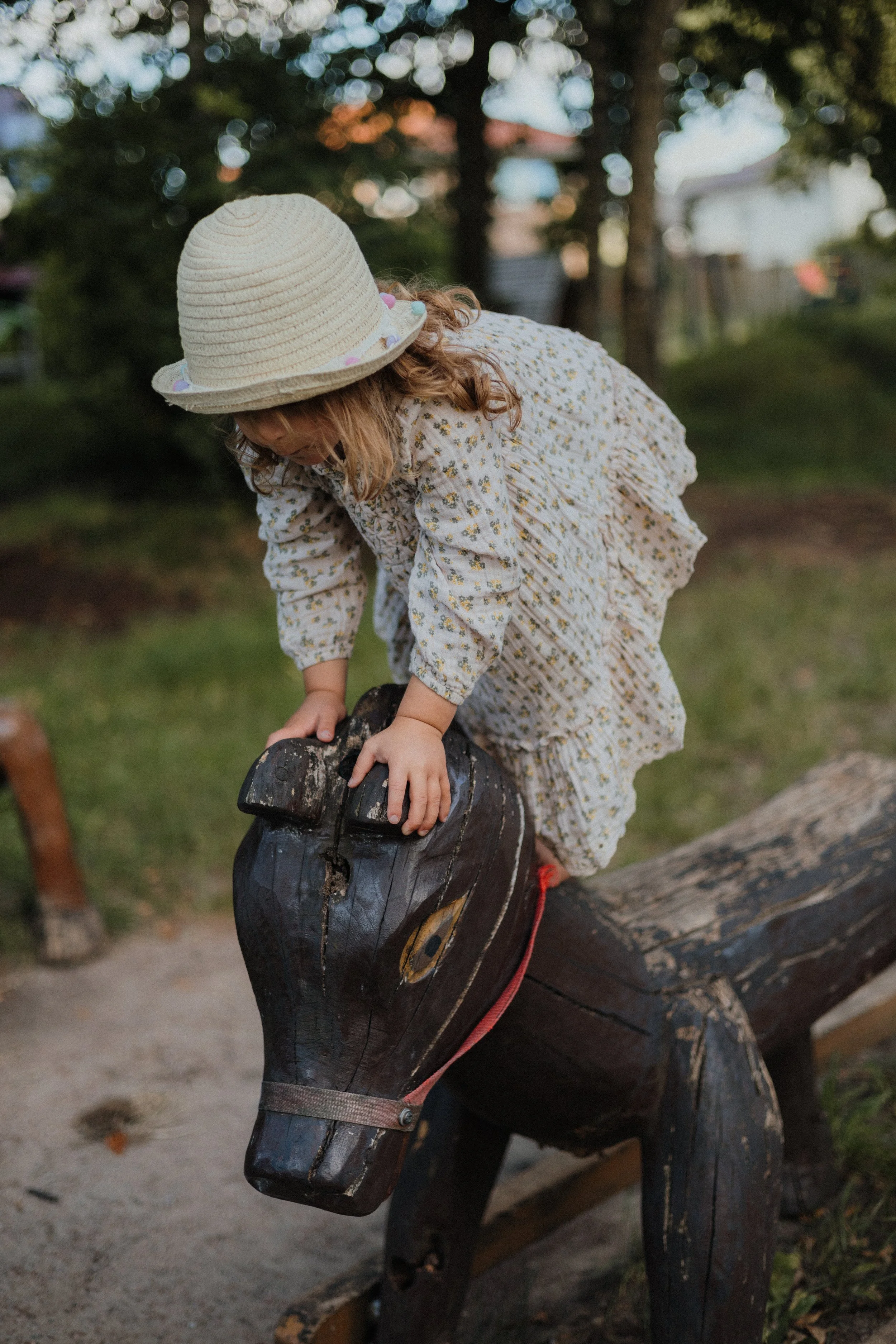 Kleines Mädchen in Blumenkleid und Strohhut, das auf einem Holzpferd im Freien sitzt.