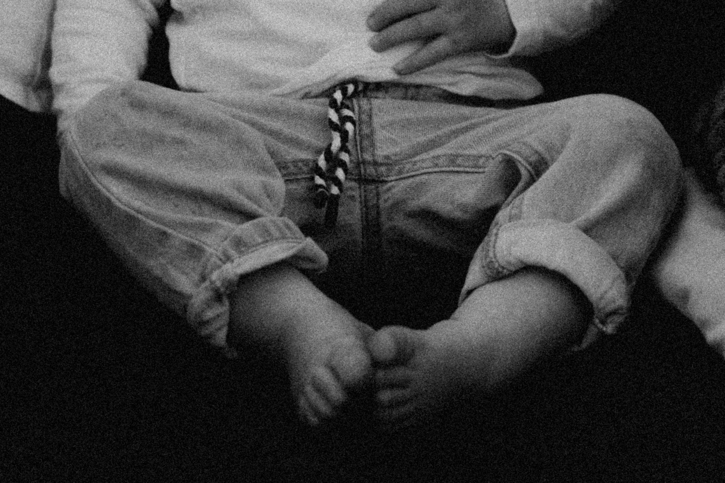 Schwarzweißaufnahme eines Kleinkindes, das auf einem dunklen Hintergrund sitzt, mit einem Ärmel hochgekrempelt und einer kuscheligen Hand in der Nähe.