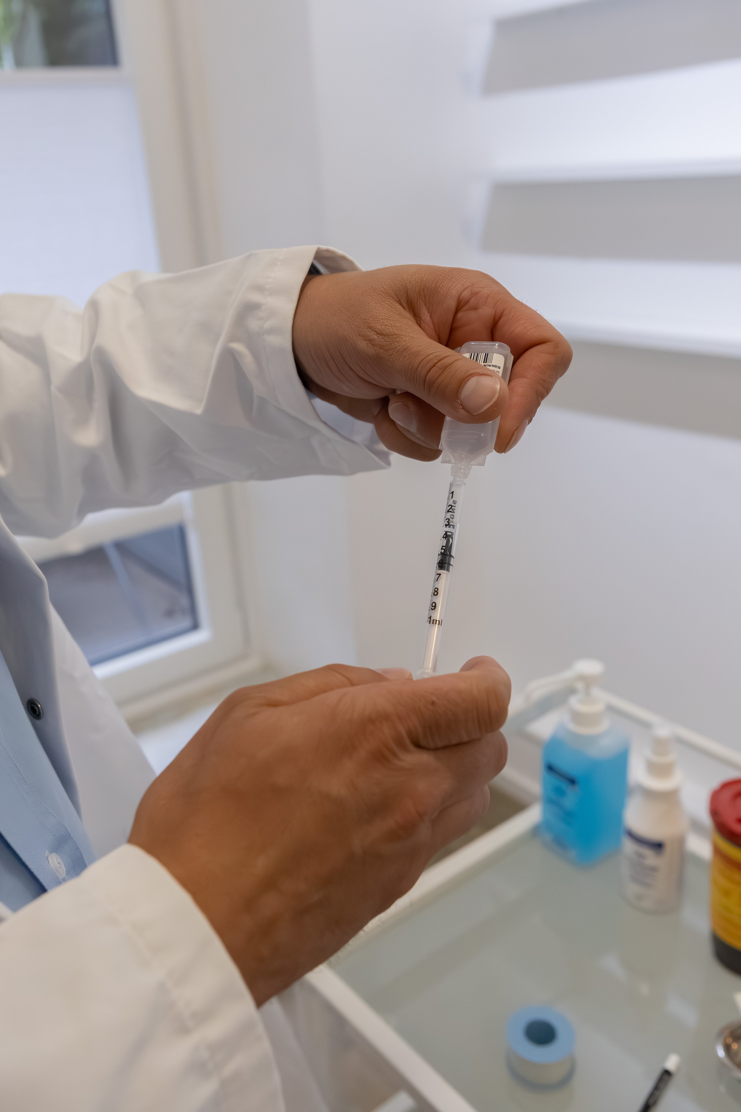 Person in einem weißen Kittel zieht eine Spritze mit Impfstoff oder Medikament vor. Im Hintergrund sind verschiedene Flaschen und Behälter auf einem Waschbecken, vermutlich in einem medizinischen Umfeld.