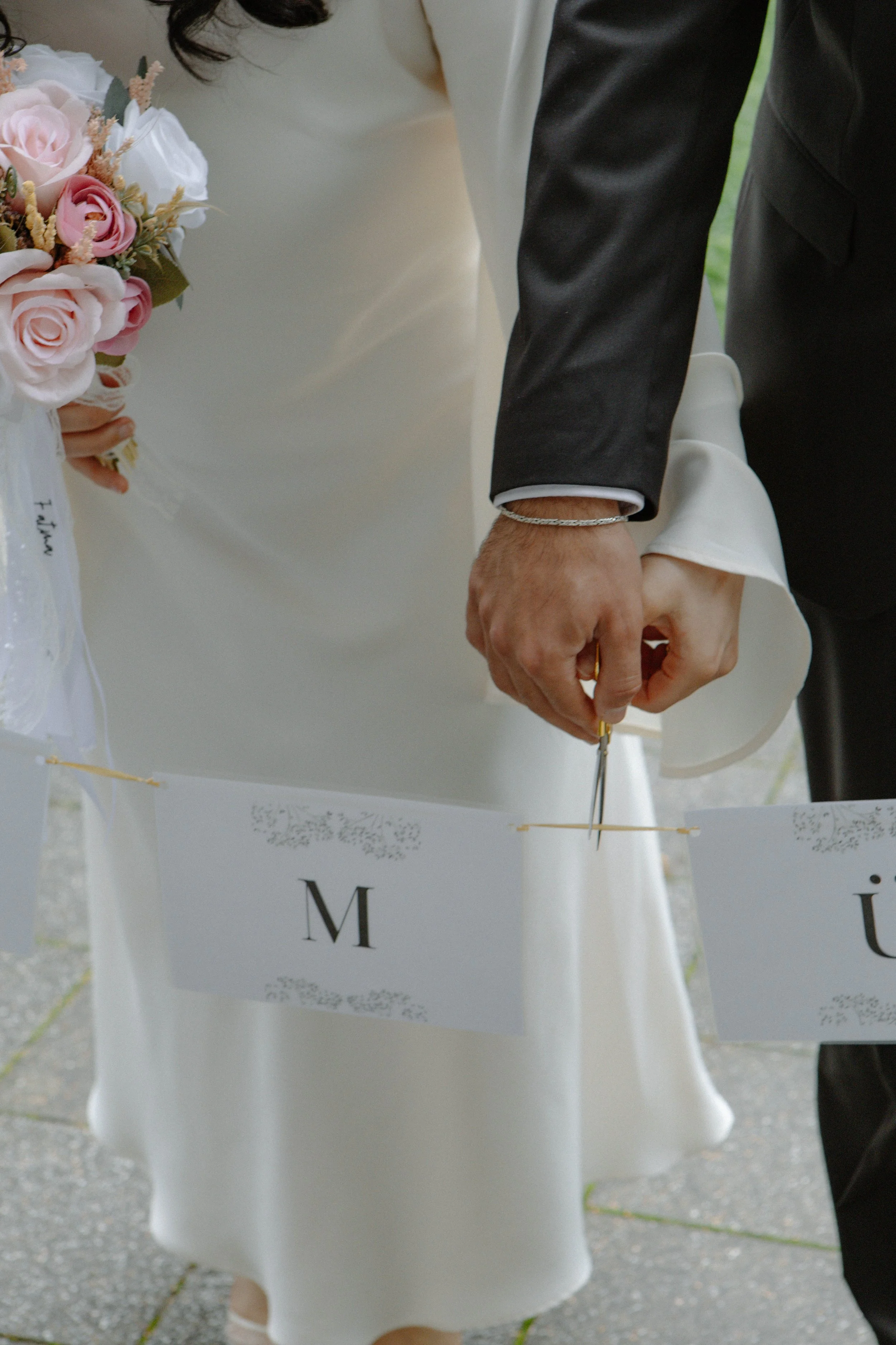 Ein Brautpaar schneidet gemeinsam eine Hochzeitstorte mit einer Schere, während sie an einem Band mit Buchstaben für die Sitzplatzzuordnung vorbeigehen.