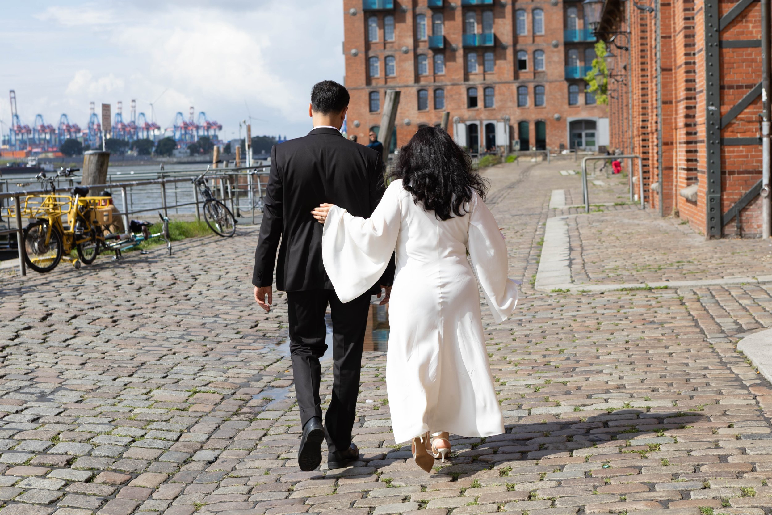 Ein Paar in formeller Kleidung, die einen Hafen entlang eines Kopfsteinpflasters gehen, mit roten Backsteinhäusern im Hintergrund.