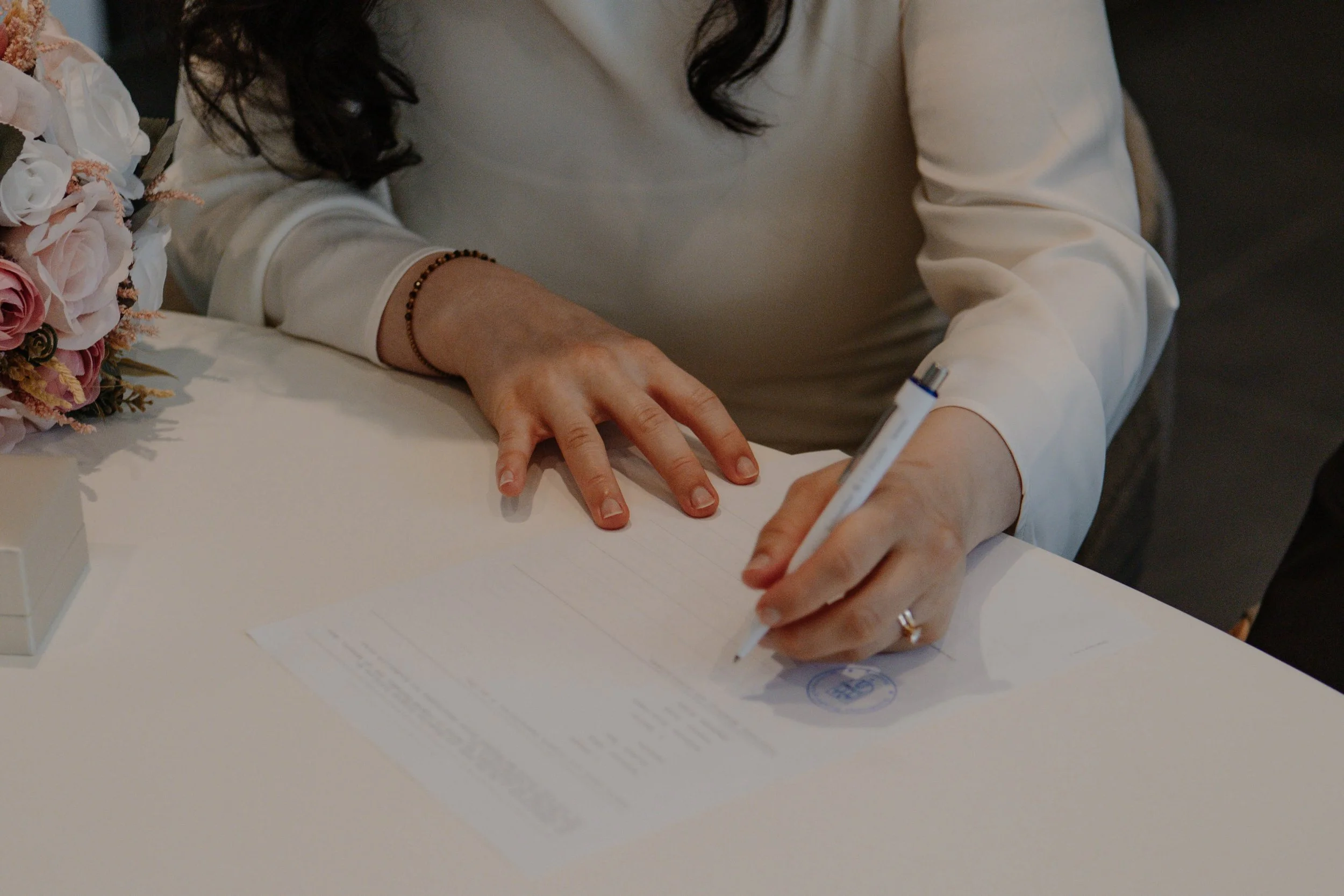 Eine Frau unterschreibt ein Dokument auf einem weißen Tisch, neben einem Blumenstrauß.