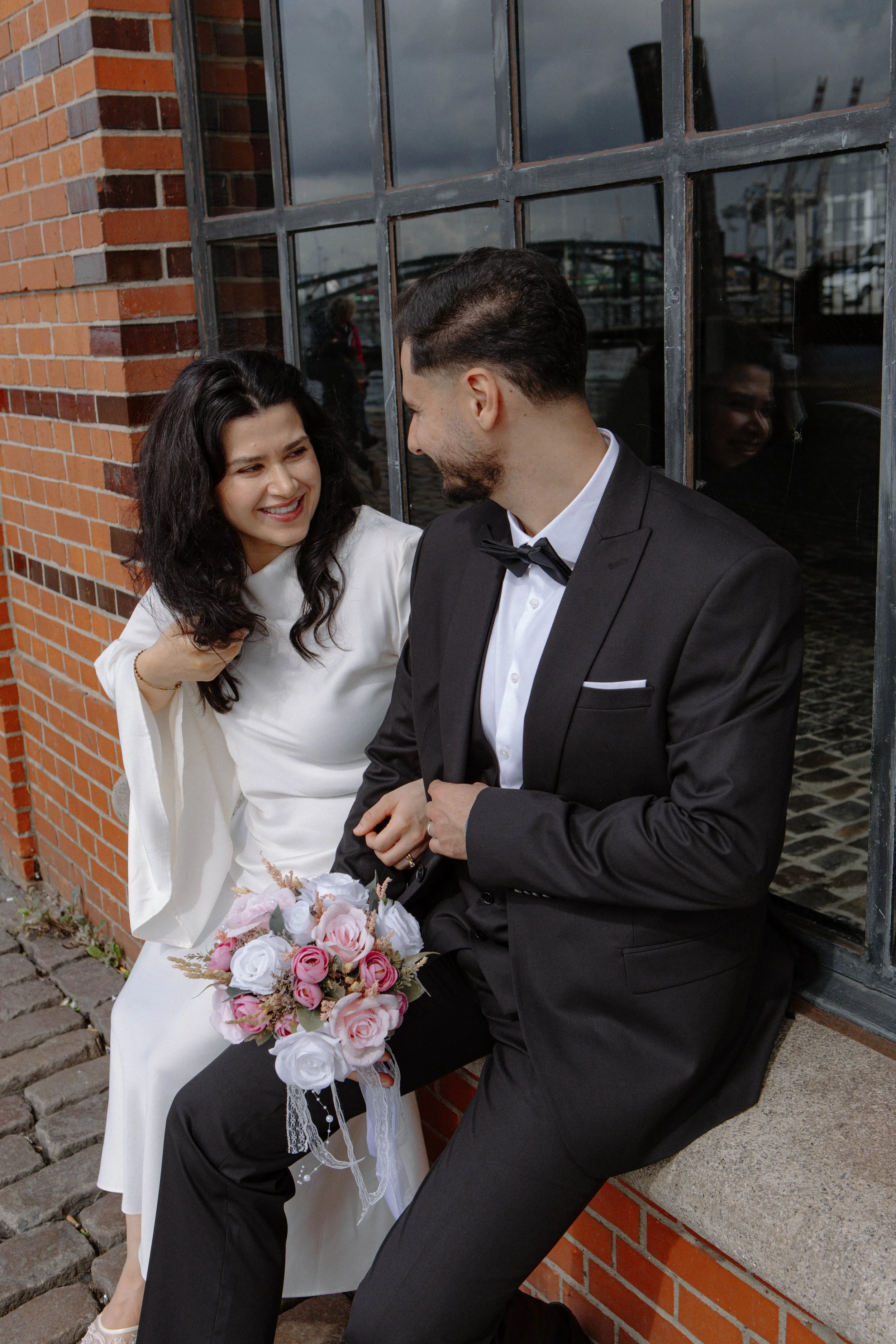 Ein lächelndes Paar auf einem Straßenrand, das an einem Tag mit Wolken am Himmel posiert. Die Frau trägt ein weißes Kleid und hält einen Blumenstrauß aus rosa und weißen Rosen. Der Mann trägt einen schwarzen Anzug mit Fliege.