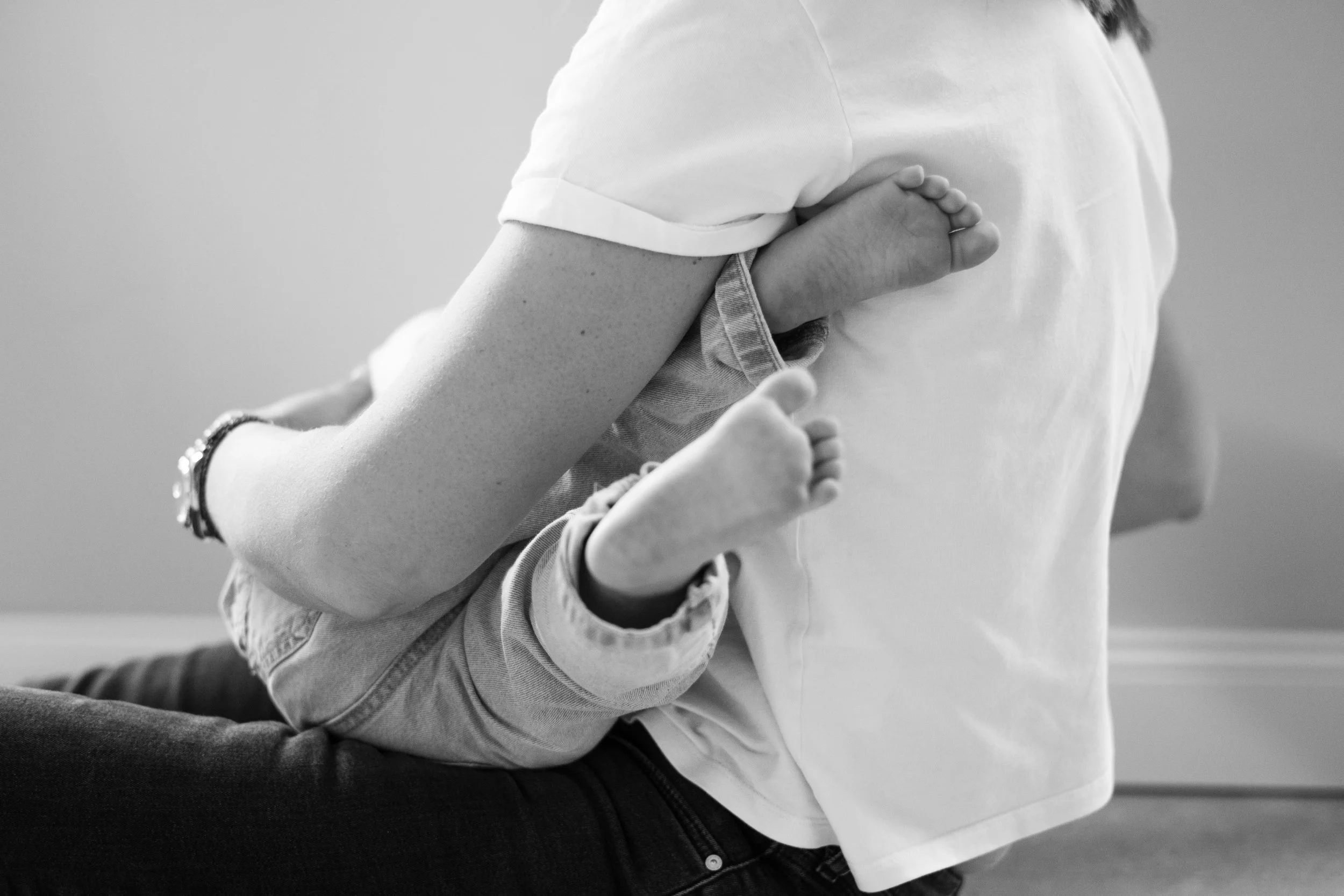 Ein Baby sitzt auf dem Schoß einer erwachsenen Person, deren Rücken im Vordergrund sichtbar ist. Das Baby trägt Jeans und ein Oberteil und liegt mit den Füßen gegen die Brust des Erwachsenen.