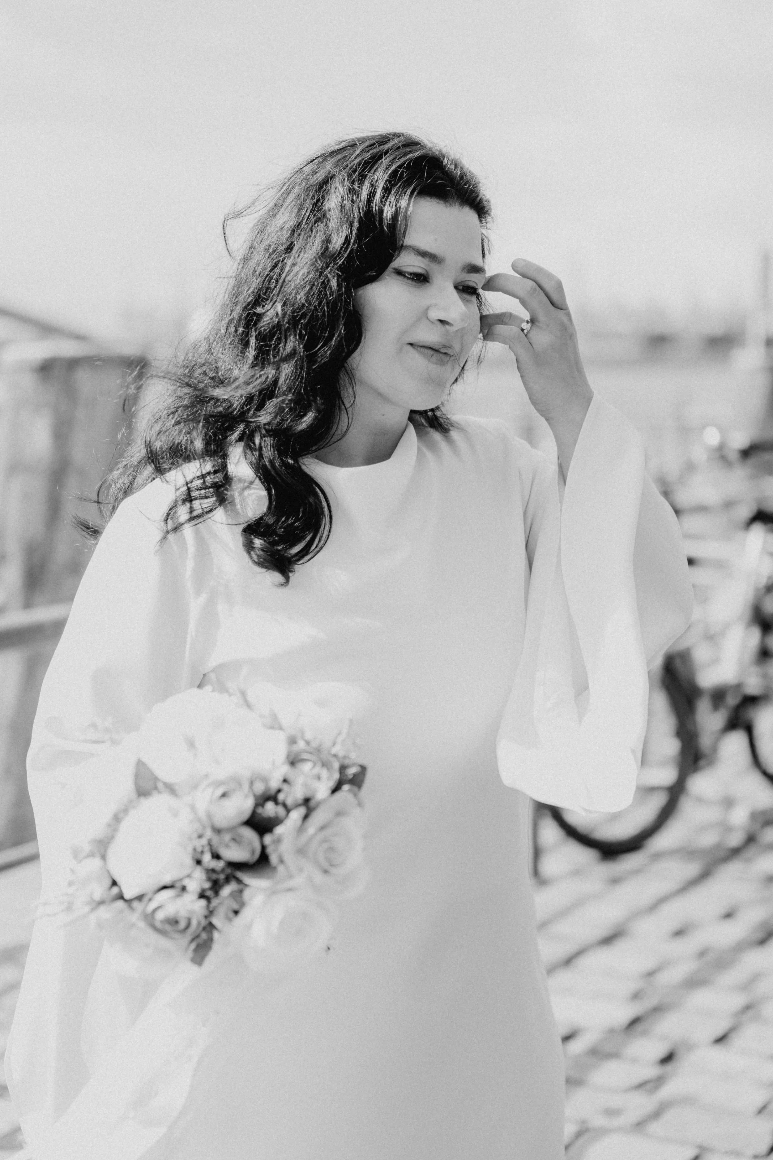 Frau mit dunklen, lockigen Haaren hält einen Blumenstrauß in der Hand und betrachtet nachdenklich ihre Frisur. Sie trägt ein langärmliges, helles Kleid.