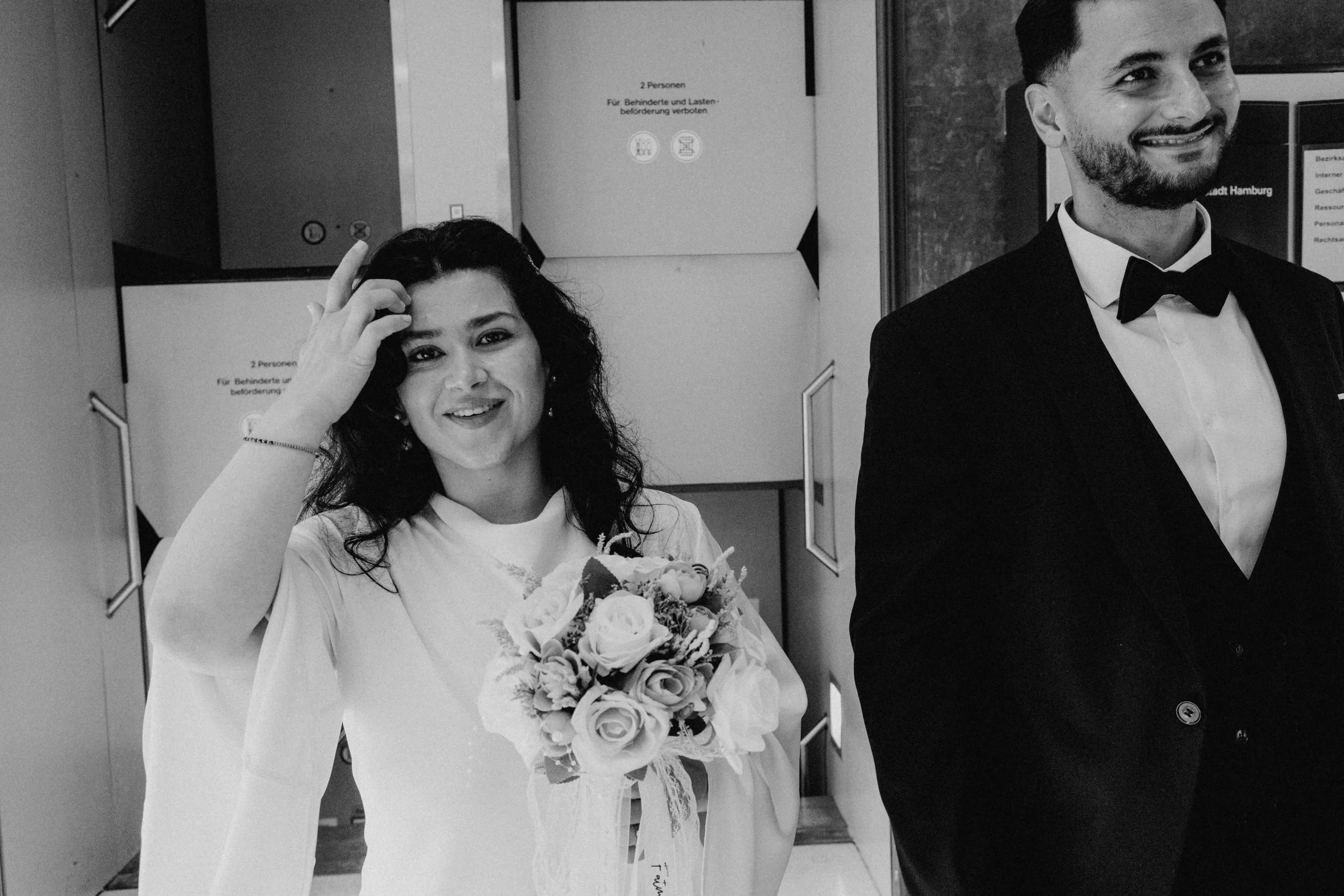 Ein lachendes Paar in formeller Kleidung, die Frau hält einen Blumenstrauß, im Hintergrund Aufzüge im öffentlichen Gebäude, schwarz-weiß Aufnahme.
