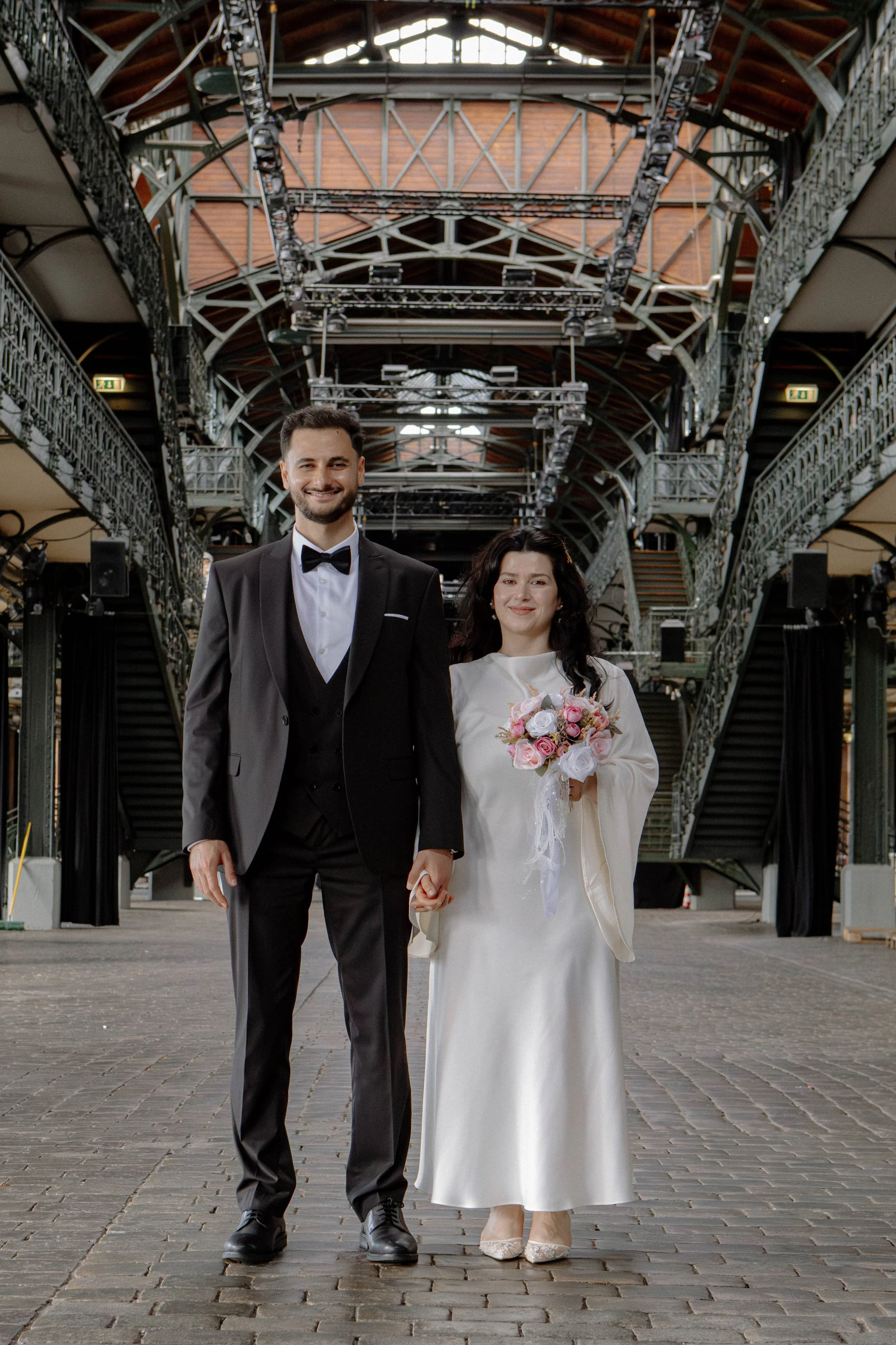 Ein verheiratetes Paar, ein Mann in einem schwarzen Anzug mit Fliege und eine Frau in einem weißen Kleid mit Blumenstrauß, steht in einem offenen, industriellen Gebäude mit hohen Decken und Treppen im Hintergrund.