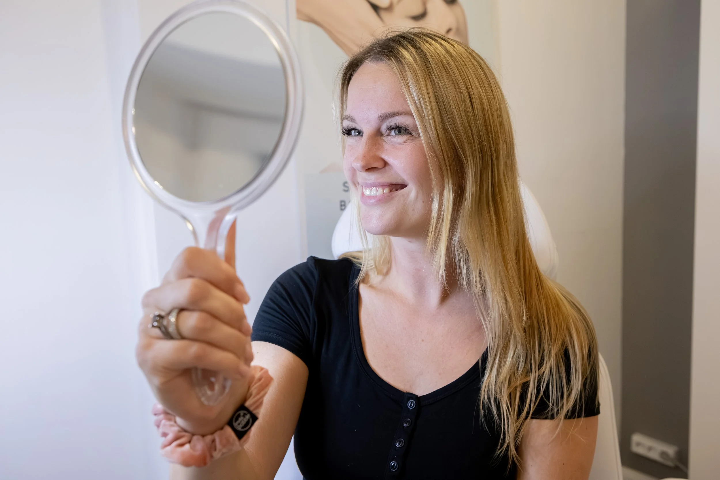 Eine lachende Frau mit blonden Haaren, die einen Handspiegel hält und sich darin ansieht.