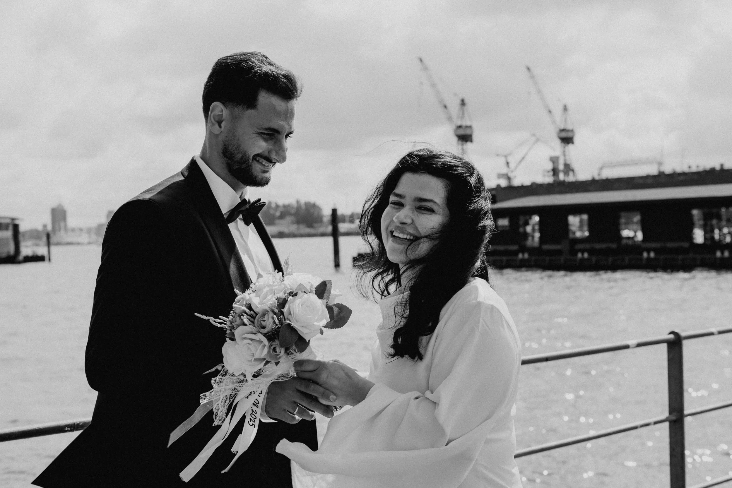Braut und Bräutigam beim Hochzeitsshooting am Wasser, lachen und halten einander Händchen, mit einer Hafen- oder Werftkulisse im Hintergrund.