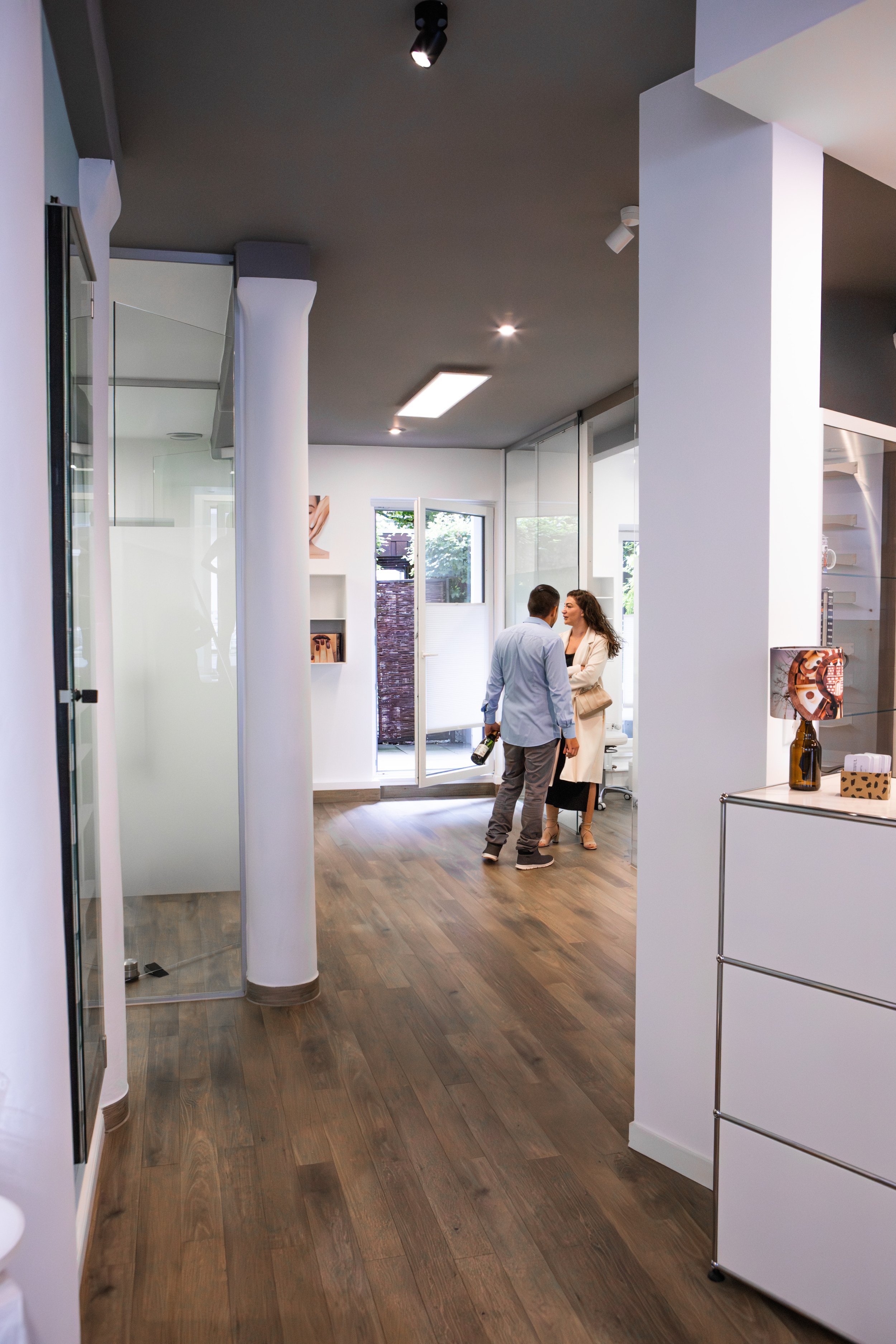 Zwei Menschen unterhalten sich in einem modernen, hellen Raum mit Holzfußboden und großen Fensterflächen.