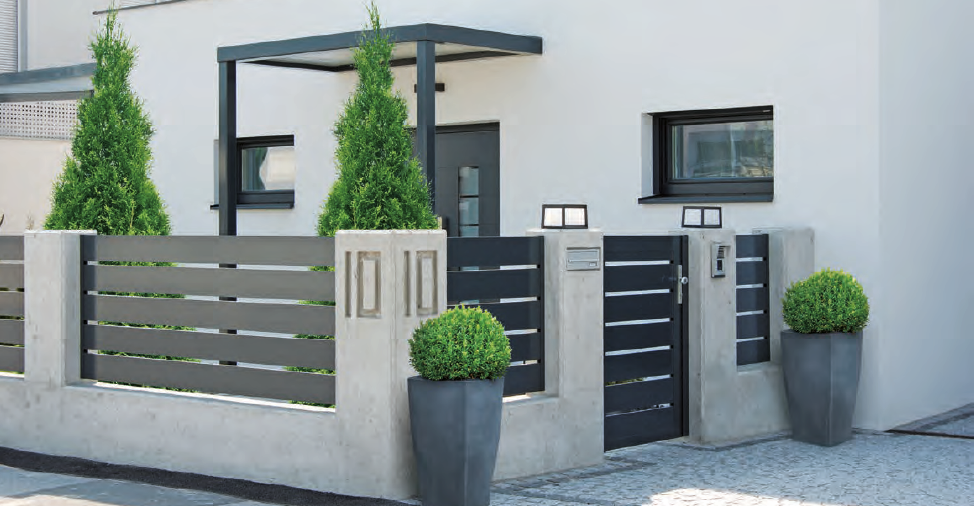 Moderne Hausfront mit weißer Fassade, schwarzem Eingangstor, zwei großen Topfpflanzen und grünen Bäumen im Vorgarten.