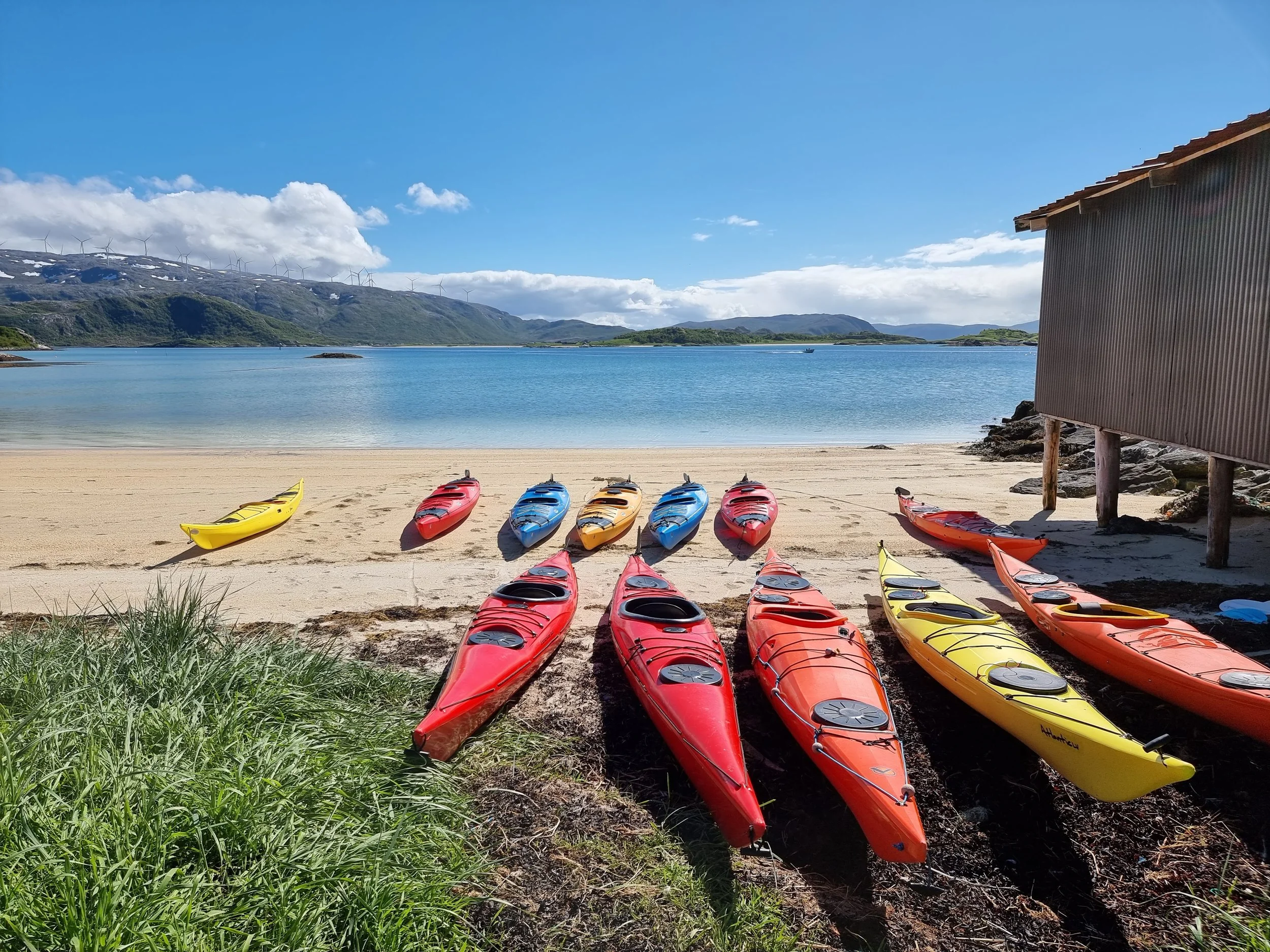 Colorful kayaks on a sandy beach, with water and mountains in the background under a partly cloudy sky.