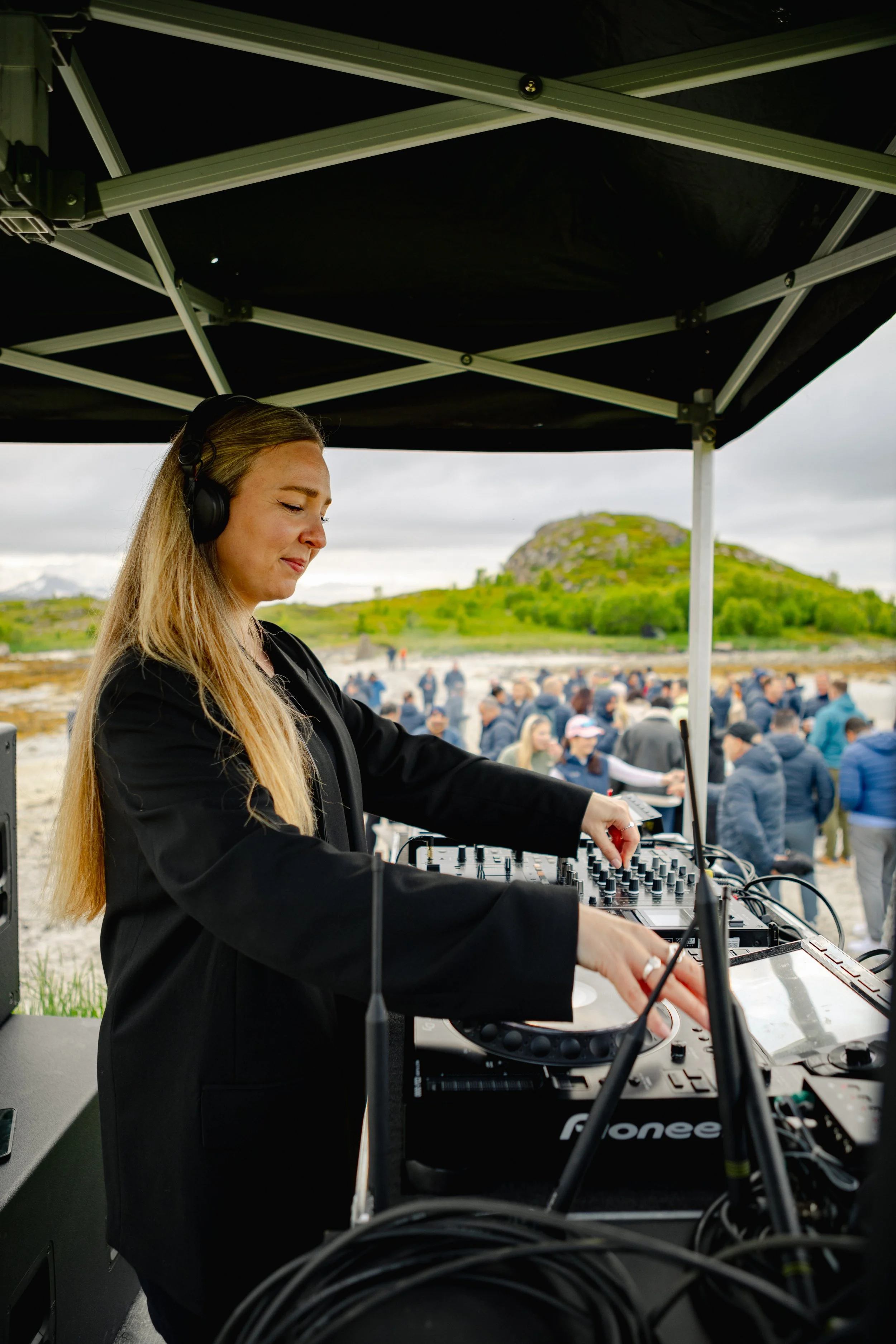 A female DJ wearing headphones performing music at an outdoor event under a black canopy, with a crowd of people and a rocky hill in the background.