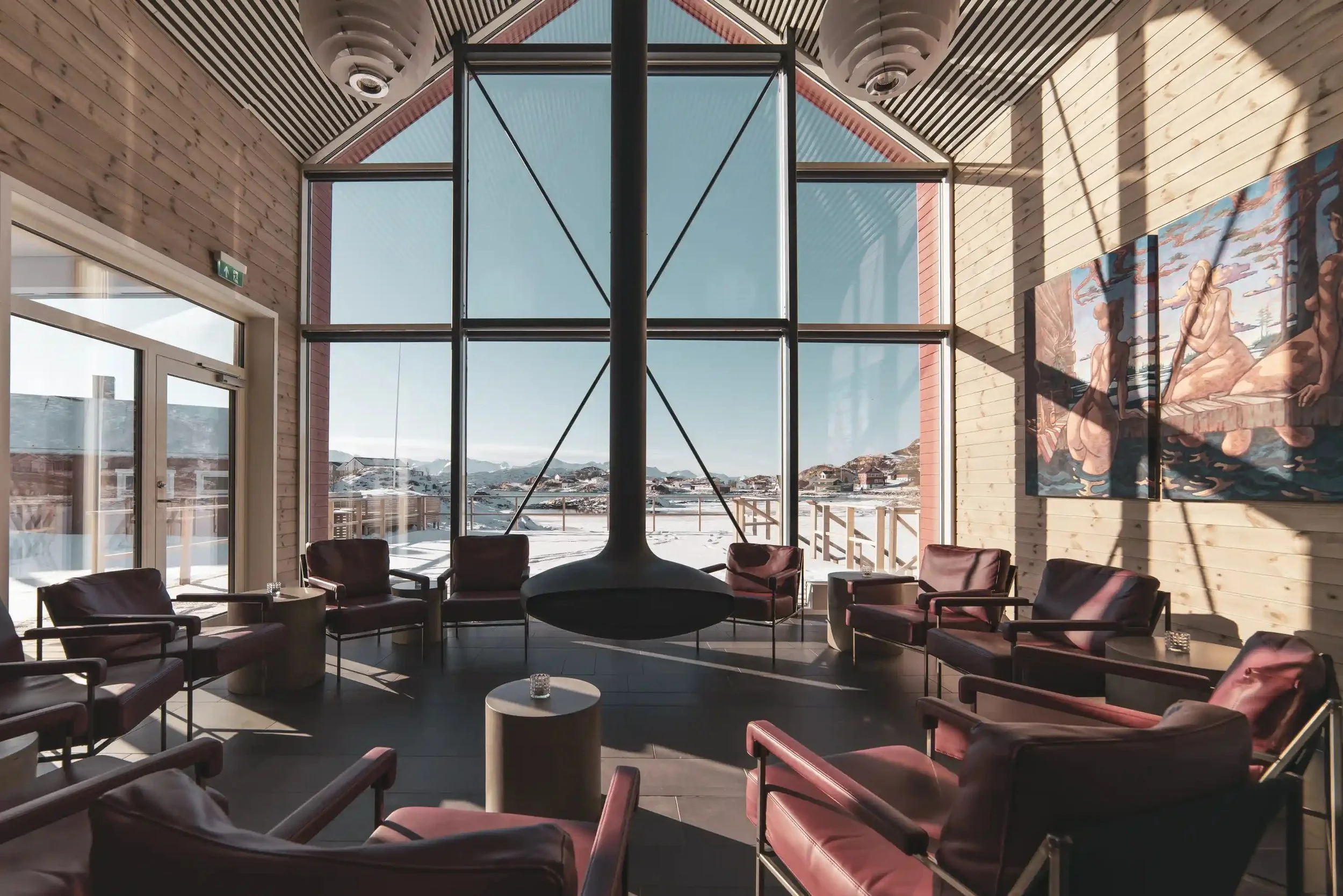 A modern lounge area with leather chairs arranged in a circle, large windows showing a snowy landscape, a central fireplace, wooden walls, and a colorful mural on the right wall.