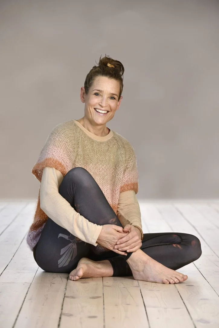 En smilende kvinde sidder på en skateboard og er klædt i et strikket oversized sweater og leggings, med en neutral baggrund.