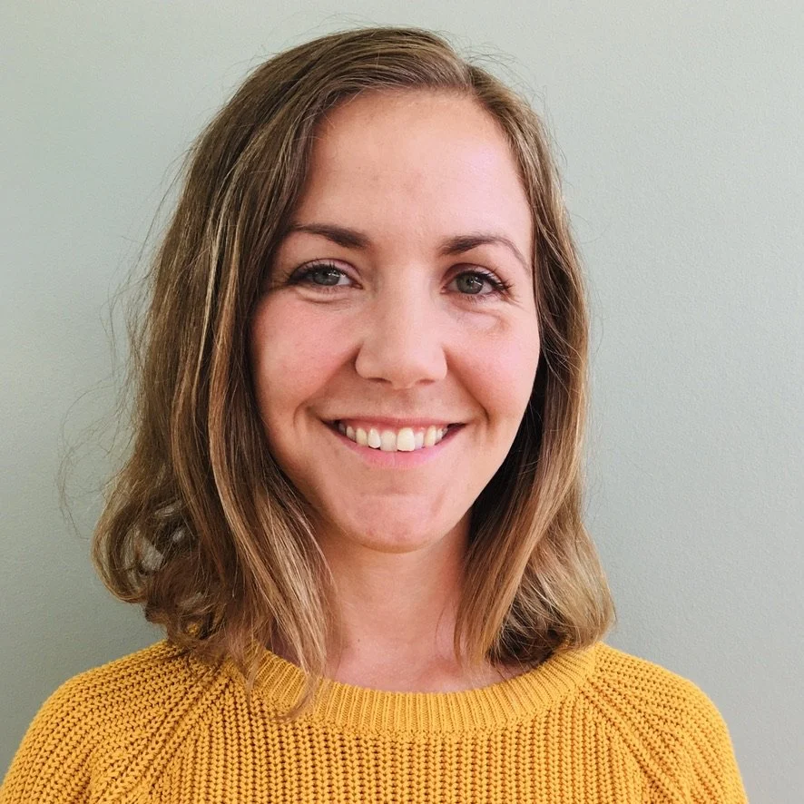 Smilende kvinde med skulderlangt, lyst hår og gul sweater stående foran en lysgrå væg.