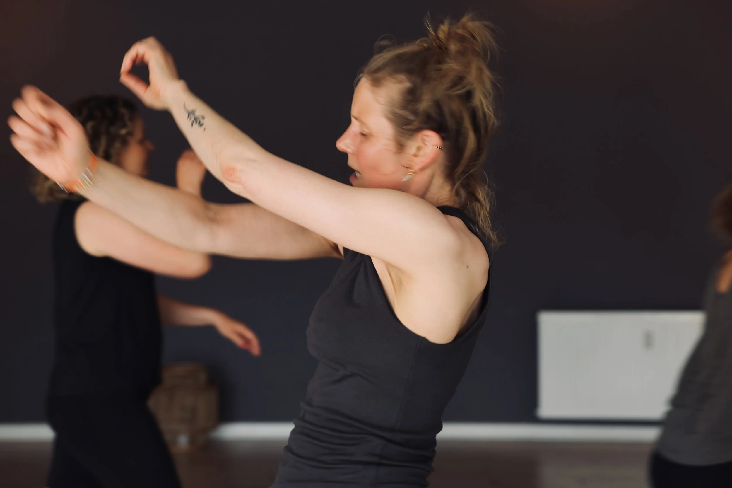 En kvinde træner eller danser i et rum med sort væg, bærer sort top, har opsat hår, og andre personer er også ude af fokus i baggrunden.