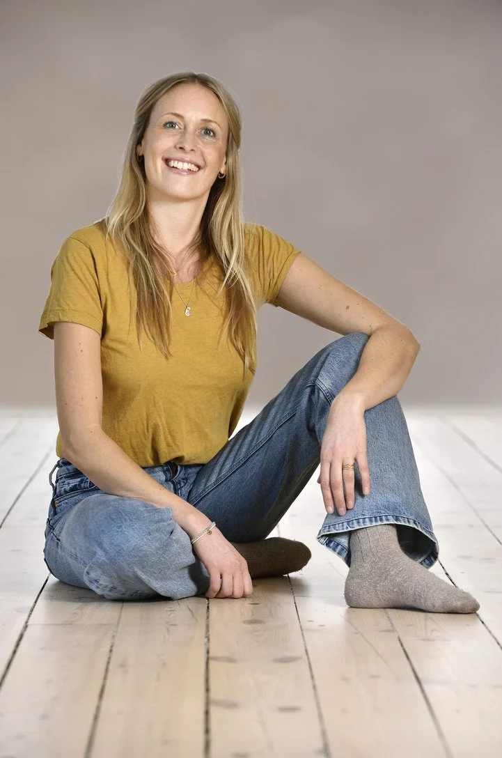 En smilende kvinde sidder på et trægulv iført en gul T-shirt, blå jeans og grå sokker, med en neutral grå baggrund.