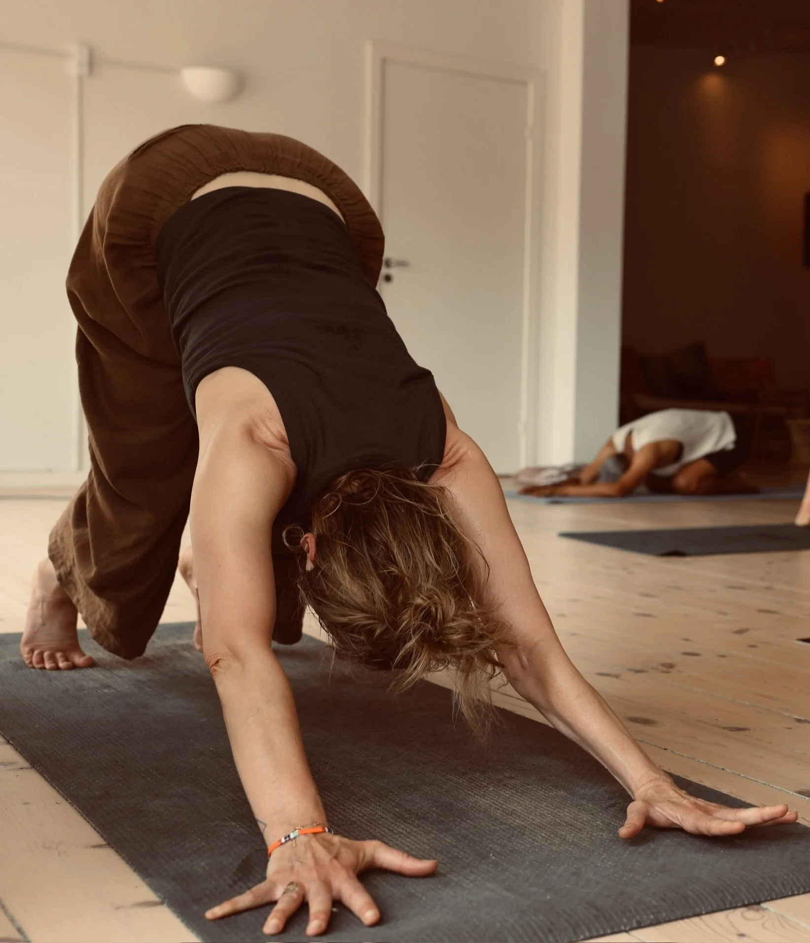 En person i yoga positur, bøjning fremover med hænder på gulvet, omgivet af yogamåtter i et rum.