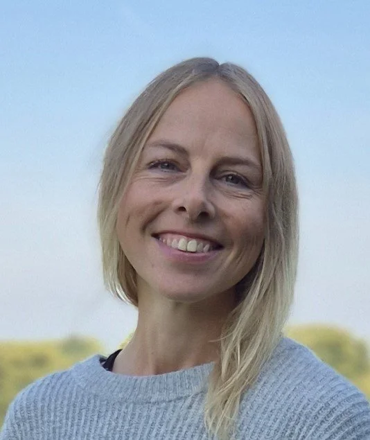 Et smilende kvindehovedbillede mod en klar blå himmel med grønne træer i baggrunden.