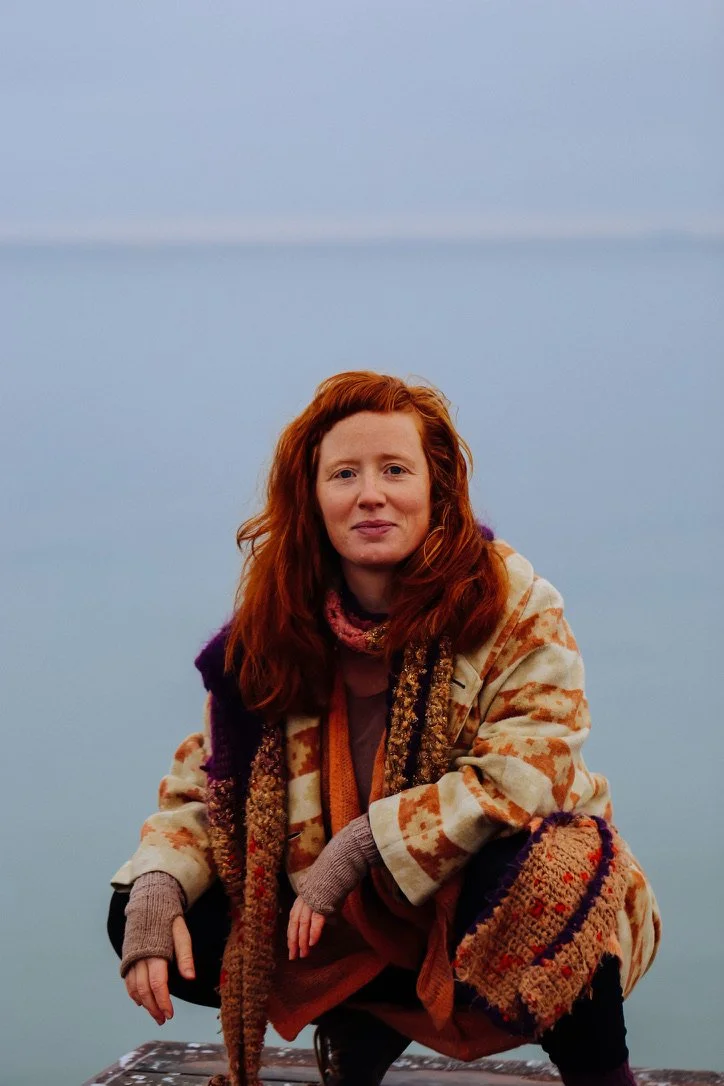 En kvinde med rødt hår iført et farverigt tøj, sidder udenfor mod en grå himmel.