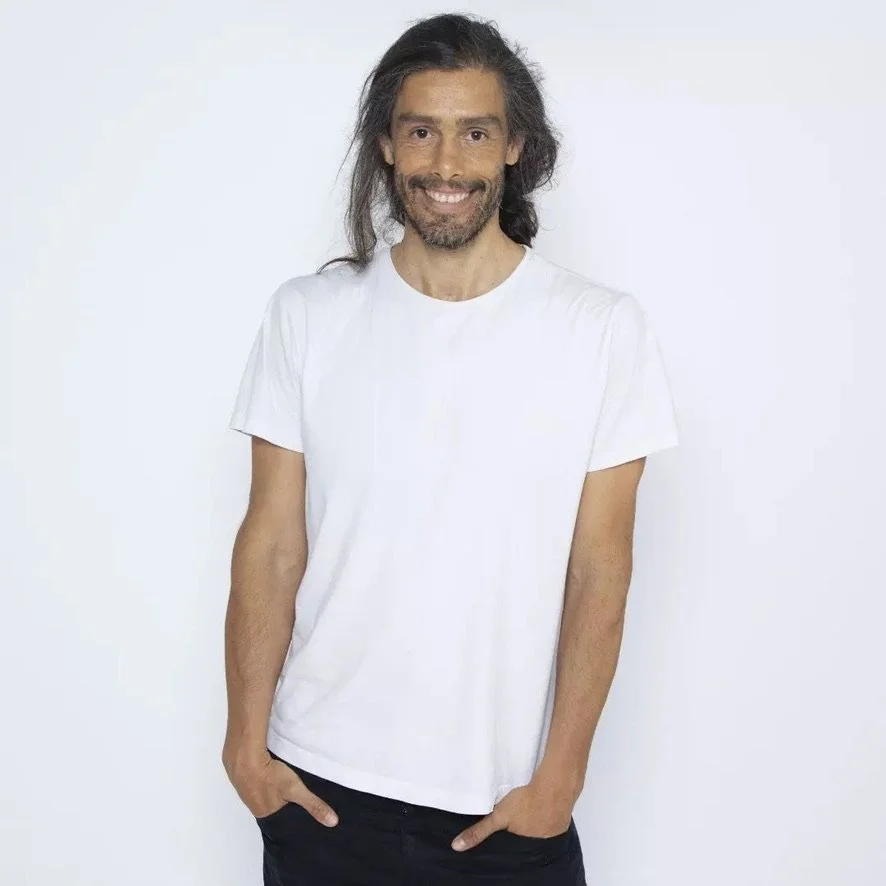 Unge mand med langt, mørkt, krøllet hår og skæg, smilende, iført hvid t-shirt og sorte bukser mod en hvid baggrund.
